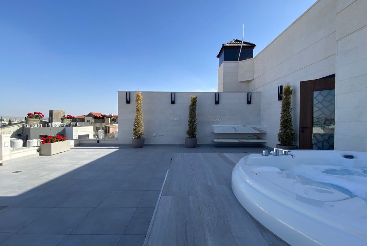 Rooftop terrace with a circular hot tub in the foreground, modern white stone walls, potted evergreens, and a distant city skyline under a clear blue sky.
