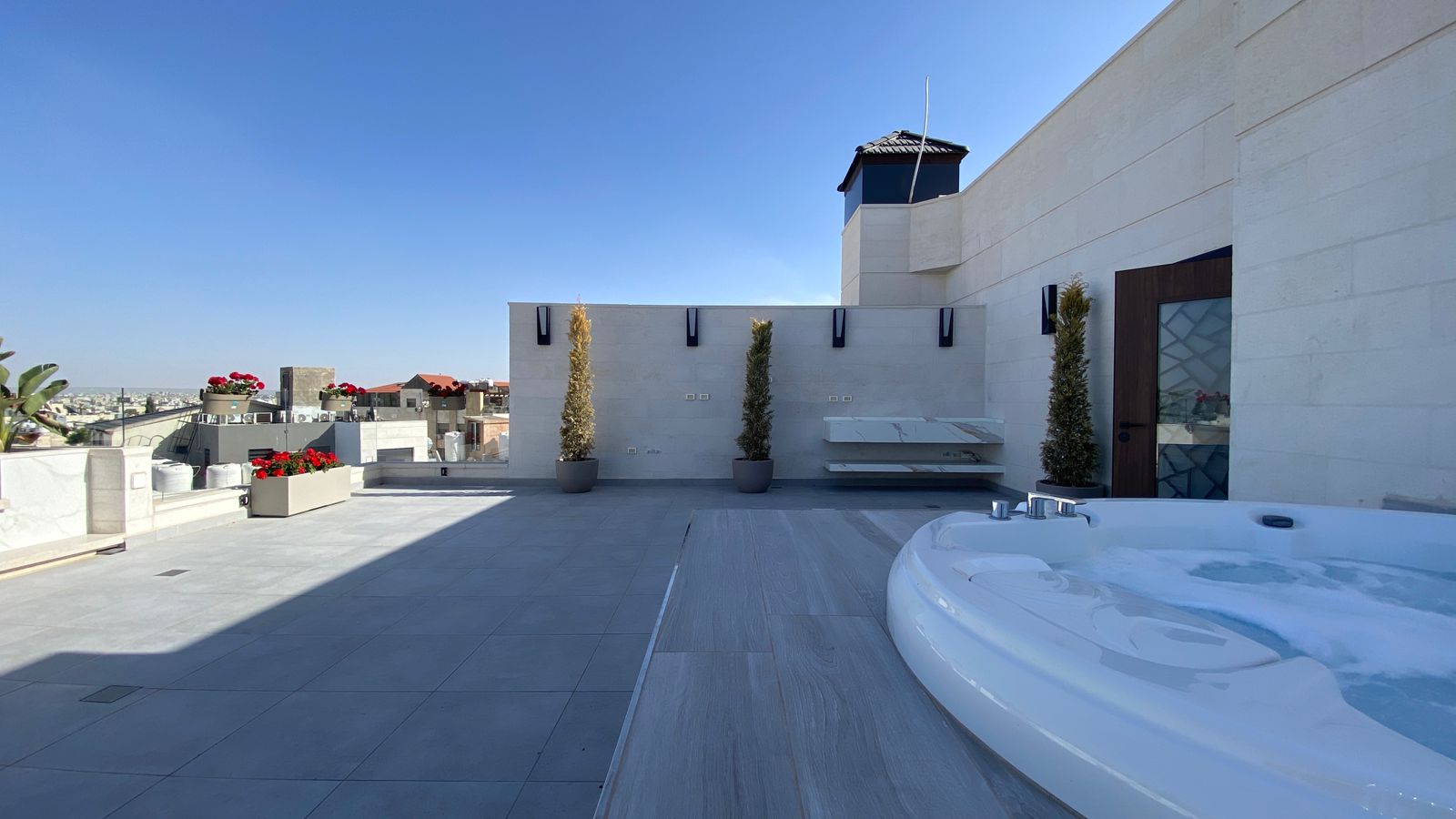 Rooftop terrace with a circular hot tub in the foreground, modern white stone walls, potted evergreens, and a distant city skyline under a clear blue sky.
