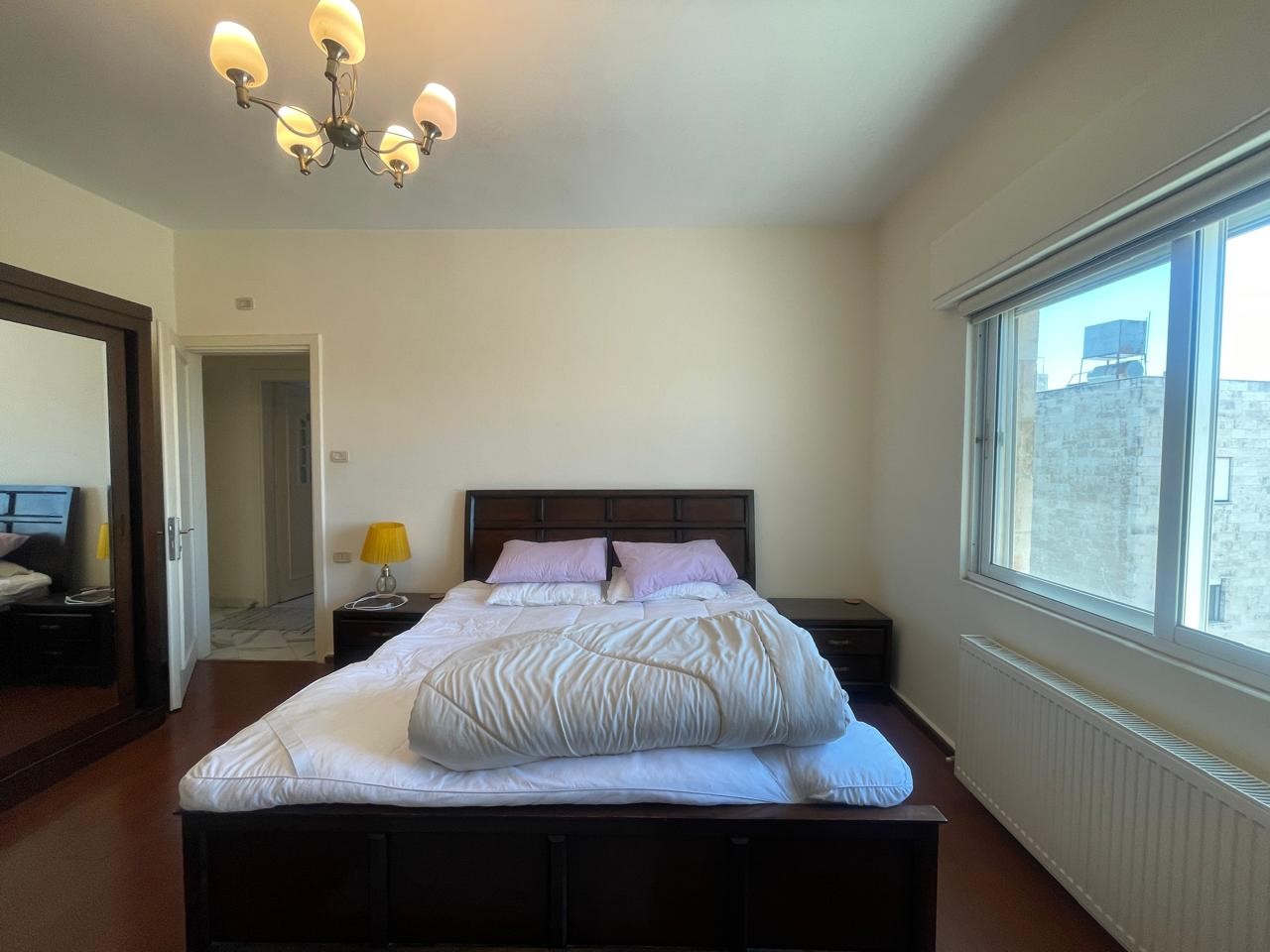 a4db15b2-8f6d-45eb-a21a-2d8e054562ba Bedroom with dark wood bed, white duvet and lavender pillows, two nightstands with yellow lamps, chandelier, and a large window letting in light.