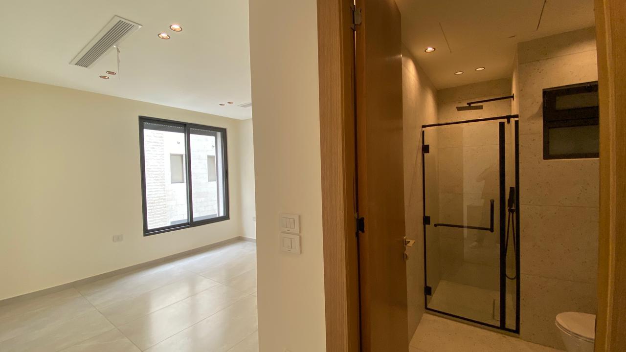 a54482d7-b8e2-4288-b20f-14104bd95ff2 Bright empty living area with a large window and light tile floor, adjacent to a glass-walled shower room in the bathroom.