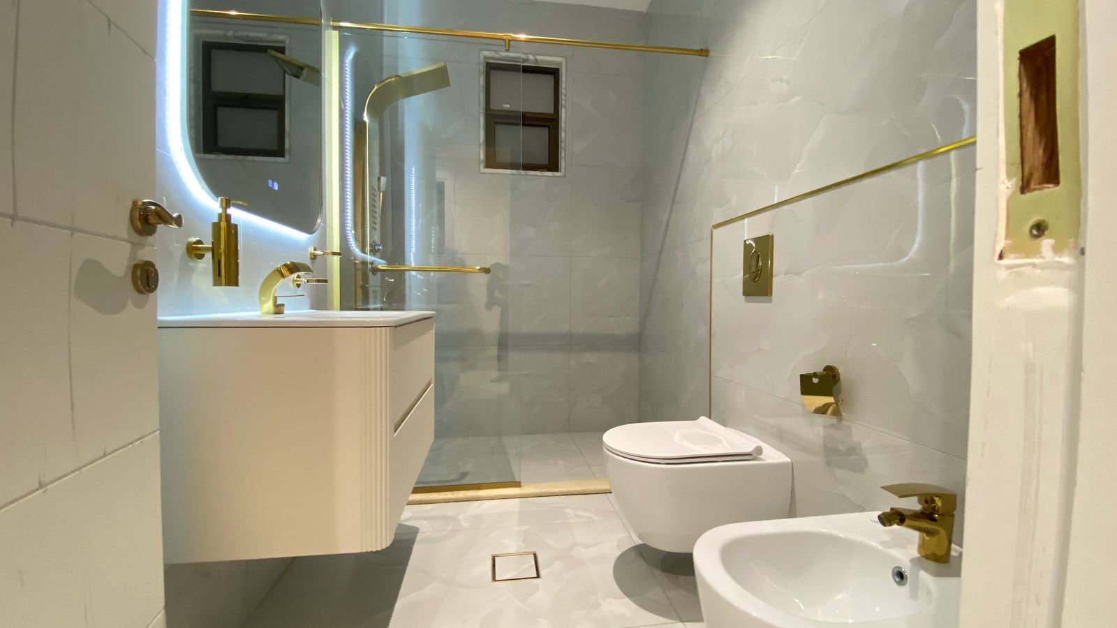 a5e29336-6a3f-4bbd-9e5c-baa715b51ea9 Bright modern bathroom with white vanity, gold fixtures, glass shower enclosure, and a wall-mounted toilet and bidet.