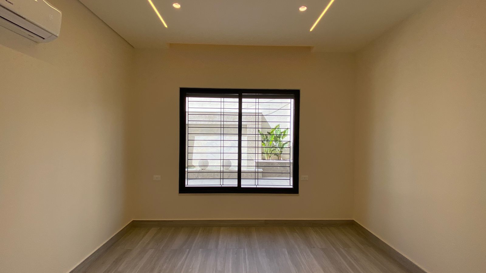 a680fdfd-2d9d-412a-a160-fc2f798cafa4 Empty rectangular room with light wood flooring, beige walls, a wall-mounted AC unit, and a barred window overlooking a small outdoor planter.