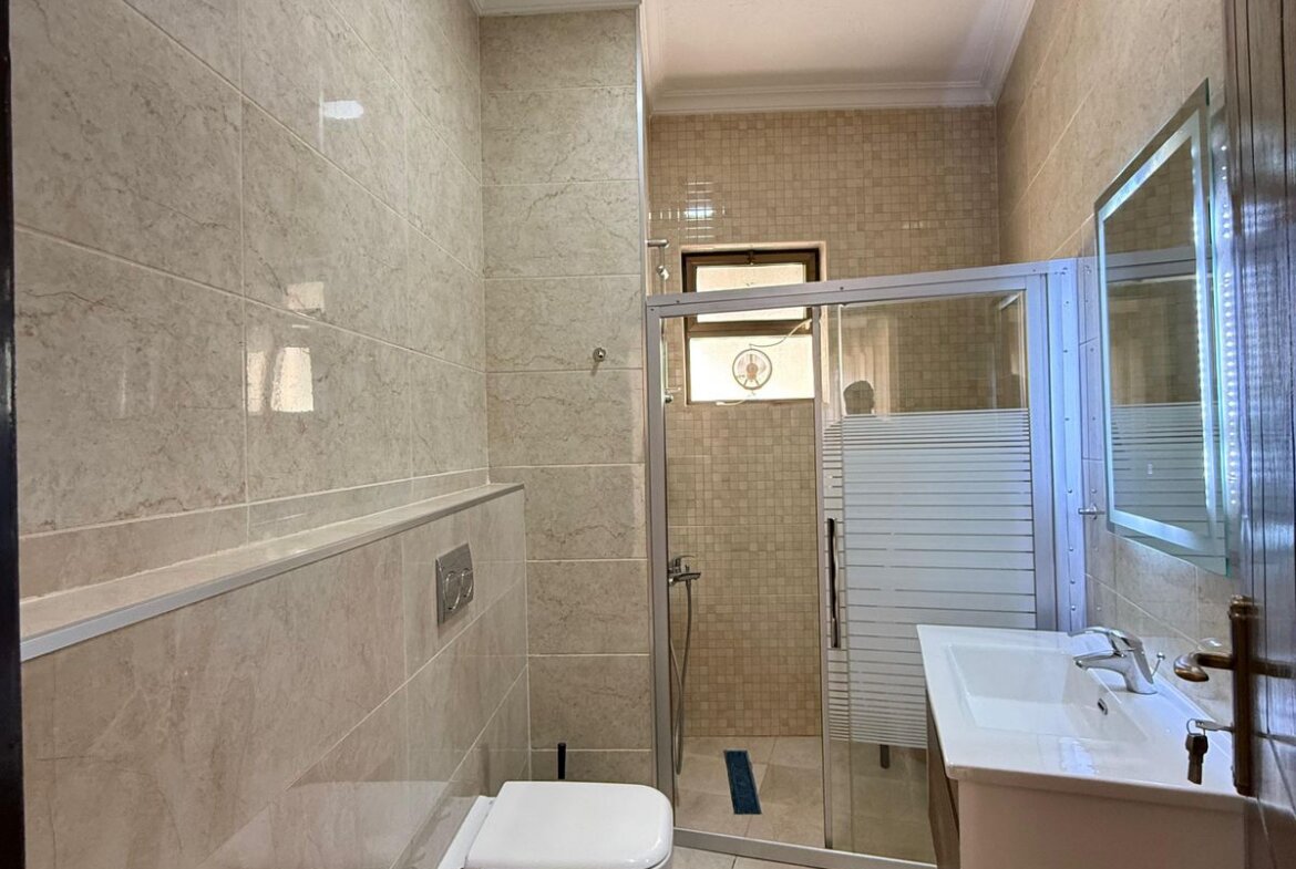 Modern beige-tiled bathroom with a toilet, bidet, white sink, and glass shower enclosure.