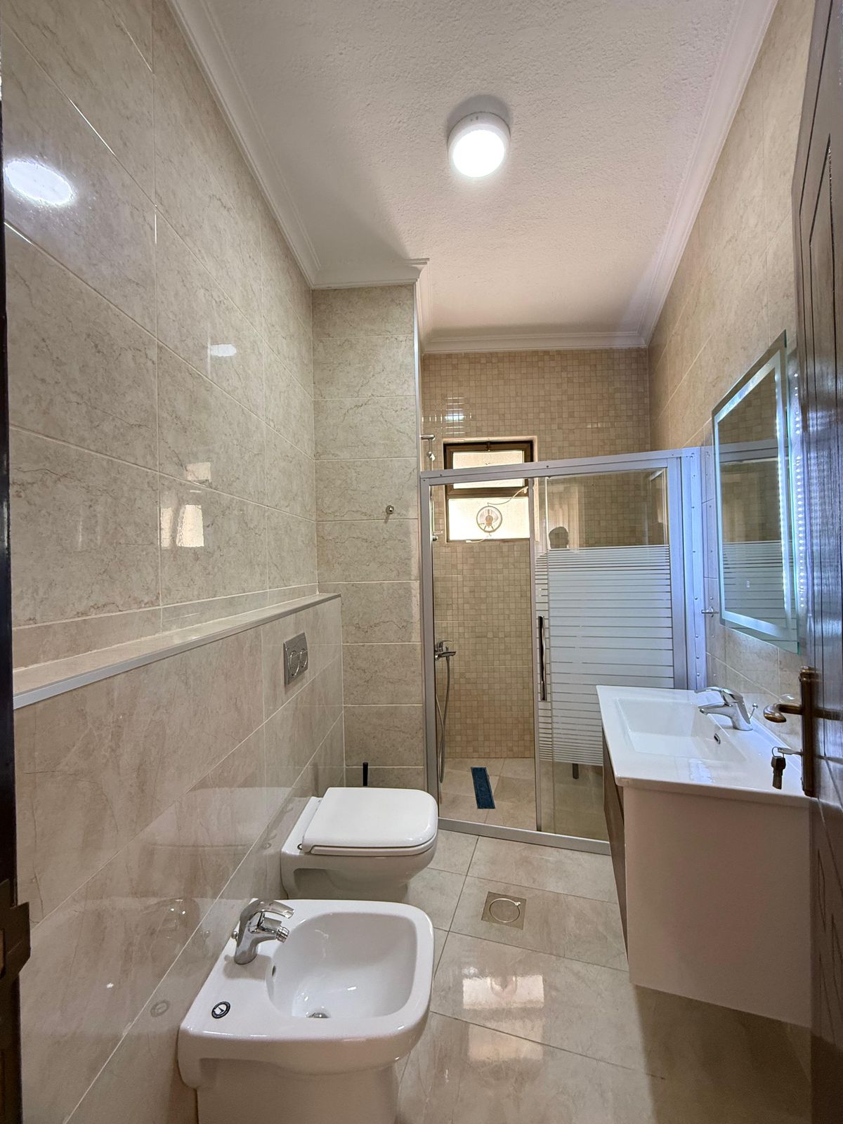 Modern beige-tiled bathroom with a toilet, bidet, white sink, and glass shower enclosure.
