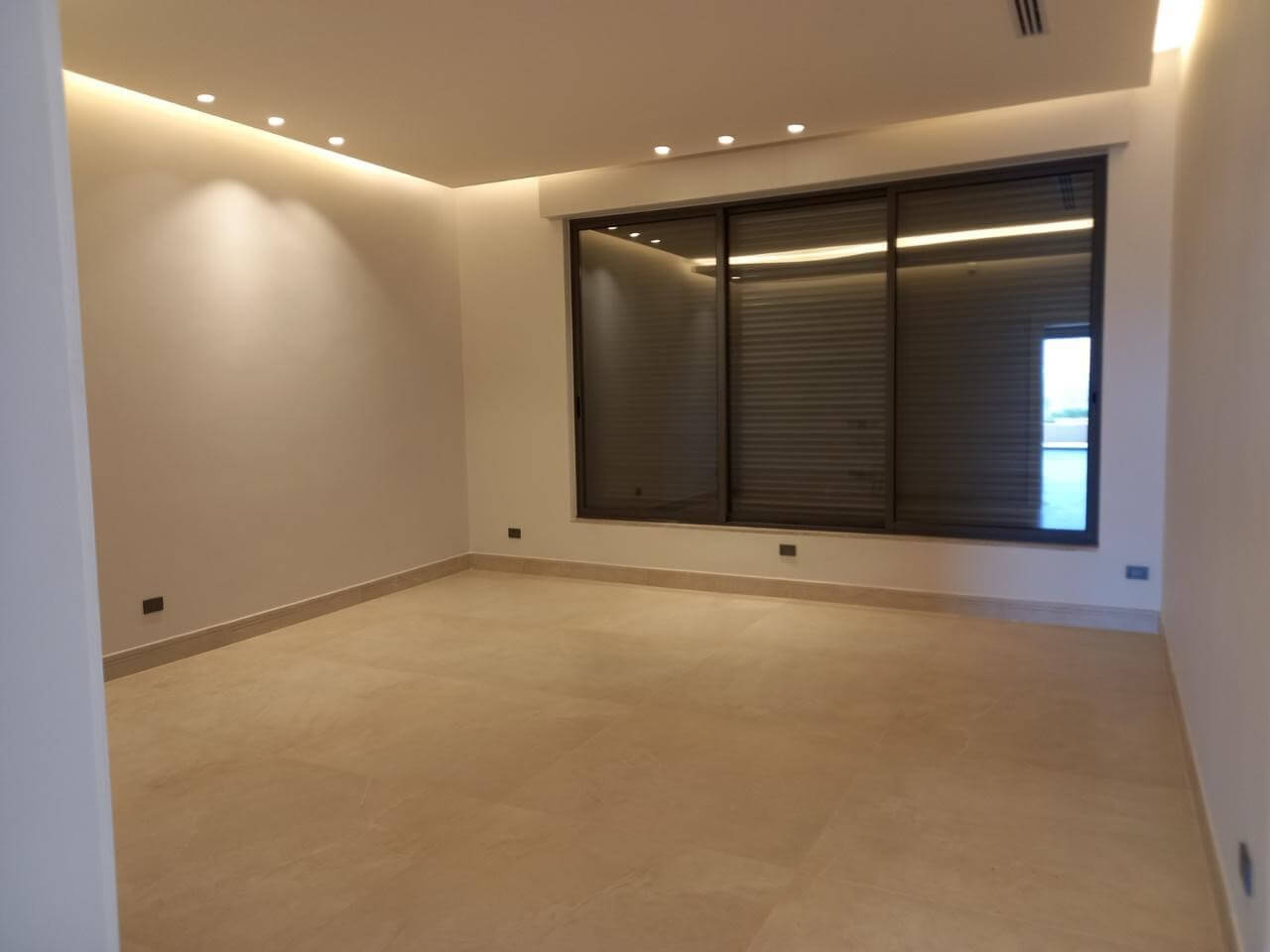 a71aa486-c872-477a-b37a-33c7336d7b4b Empty modern living room with beige tile floor, neutral walls, and a large window with closed blinds