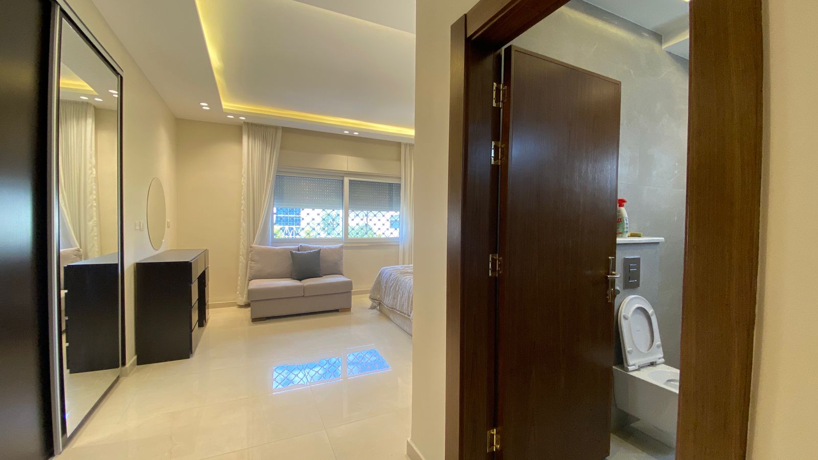 a74997a9-4498-49f1-b847-20cbbcdc8d0b Bright modern studio with cream sofa, bed, dresser, and large window; open doorway reveals a toilet in a contemporary bathroom.
