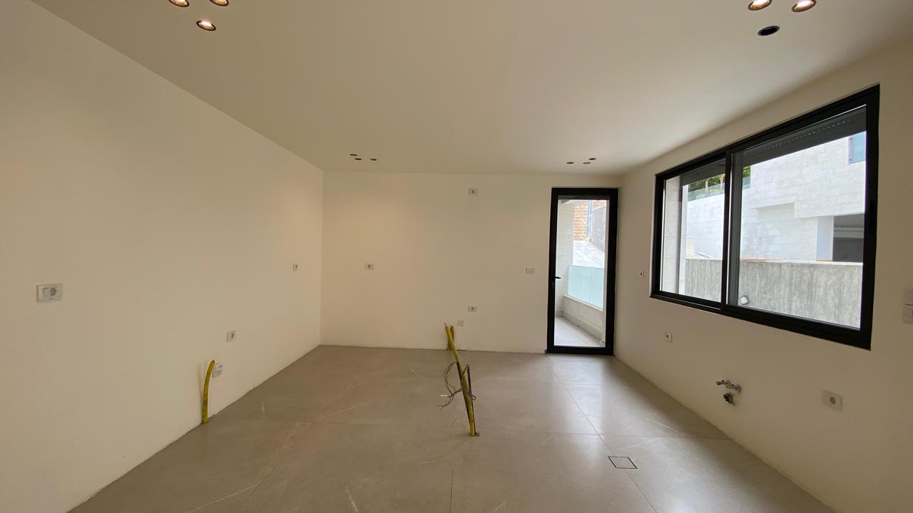 a7d4262e-1f59-498c-8ef3-1ac3c5b248b4 Empty unfinished living room with white walls, gray tile floor, ceiling spotlights, a balcony door, large window, and exposed yellow wiring.