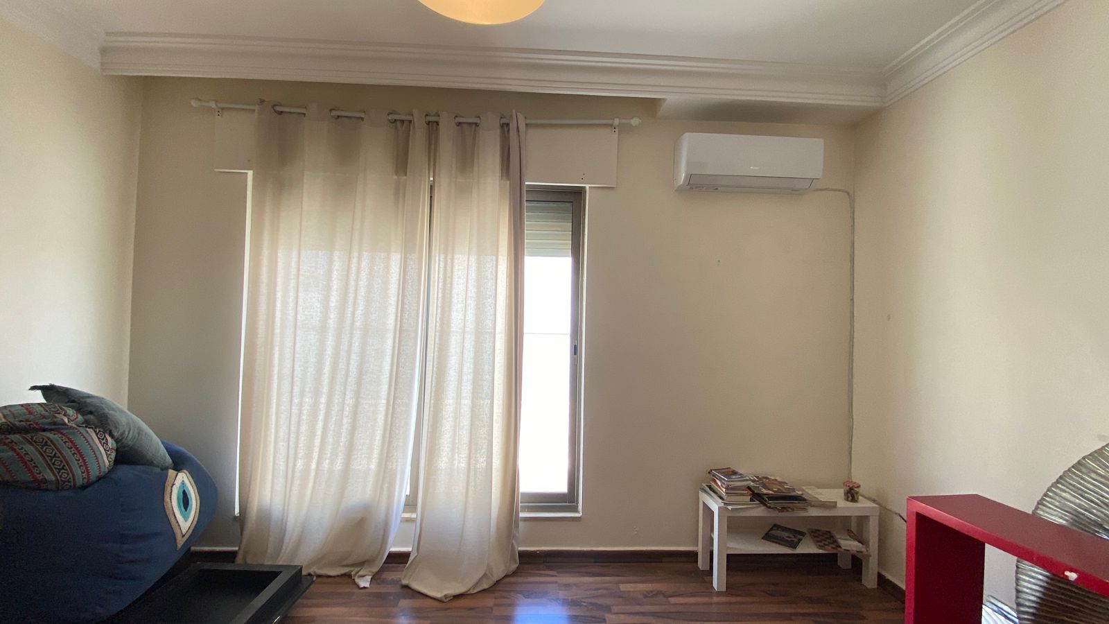 a7f4d905-46b5-4f27-b962-5806bd746ea1 Living room corner with sheer white curtains over a narrow window, a wall-mounted air conditioner, and a small white table with magazines on the right shoulder of the room.