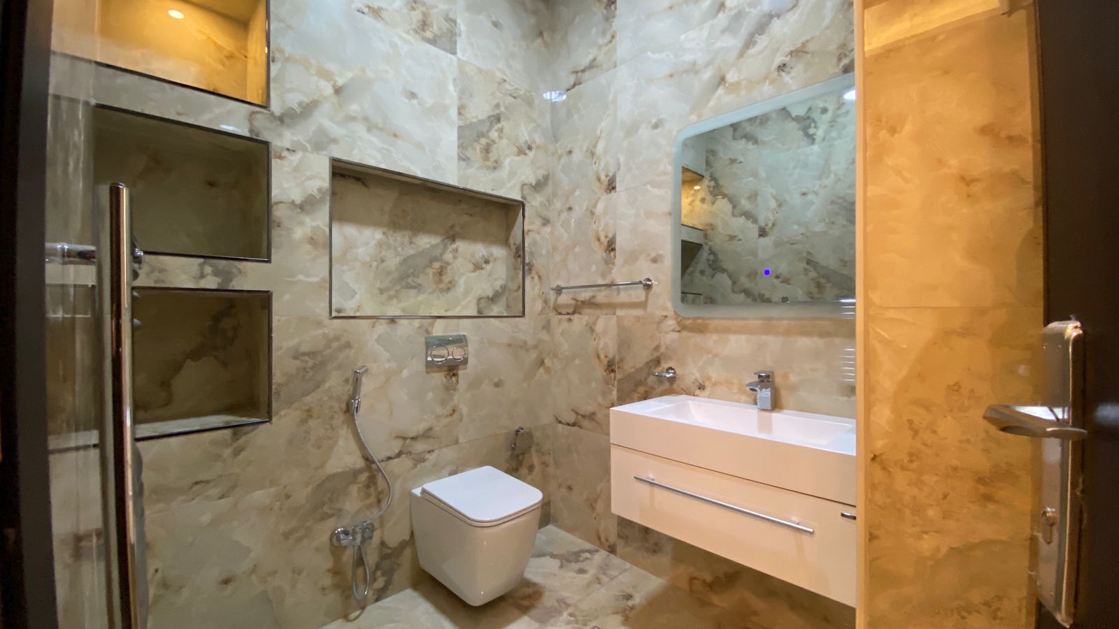 Modern marble bathroom with wall-mounted sink, floating drawers, and a wall-hung toilet; glass shower on left and large mirror above the sink.