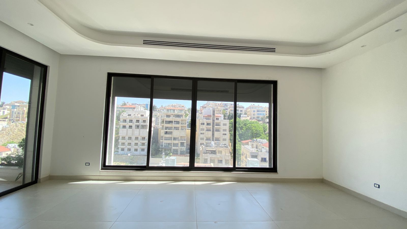 a84ba7ce-805a-4dcb-aaea-00457e6d7904 Empty white, unfurnished room with large floor‑to‑ceiling windows showing a cityscape and light tiled floor.