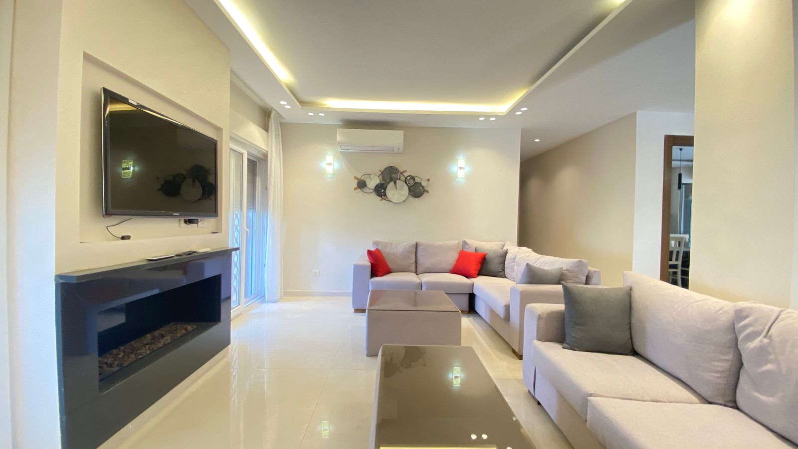 a88ed7ac-d6f0-4d7a-b06d-a03aa4c93a46 Modern living room with an L-shaped gray sofa, red and gray cushions, and a glass coffee table on a glossy floor.