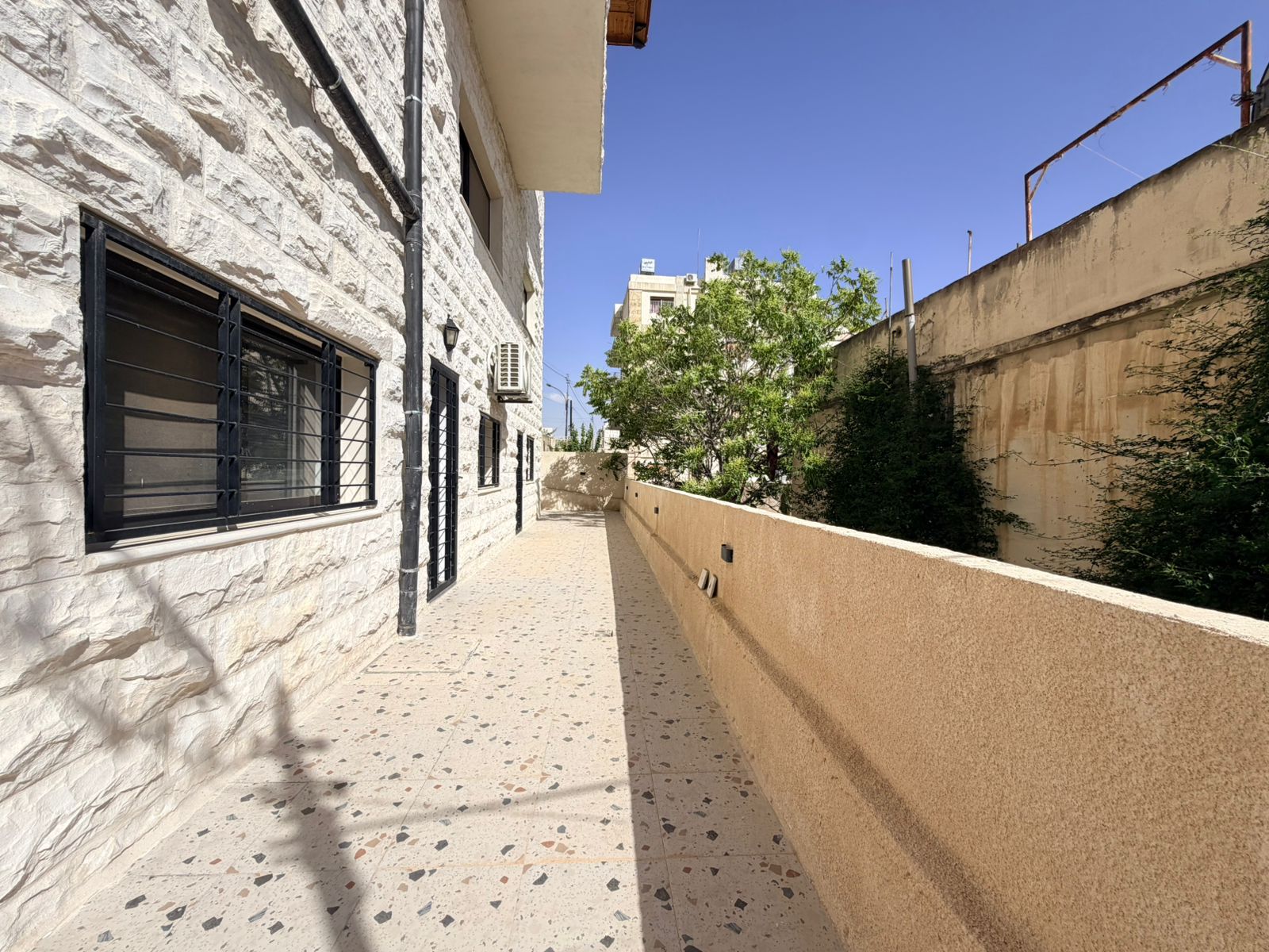 a92bac78-b3a1-4c84-b50f-749f892eb98b Sunlit narrow walkway along a white stone building with barred windows, terrazzo floor and a beige wall on the right, with trees and blue sky beyond.