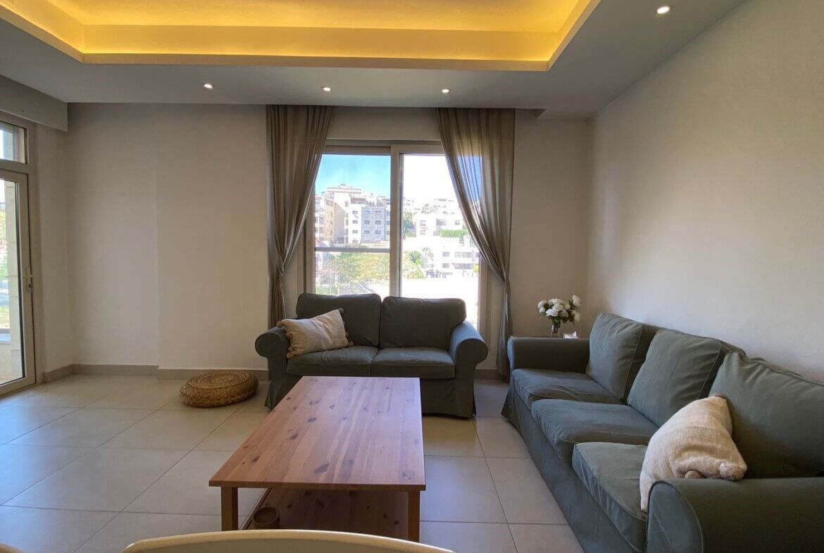 Living room with two gray sofas, a wooden coffee table, and a TV on the left; large windows with beige curtains.
