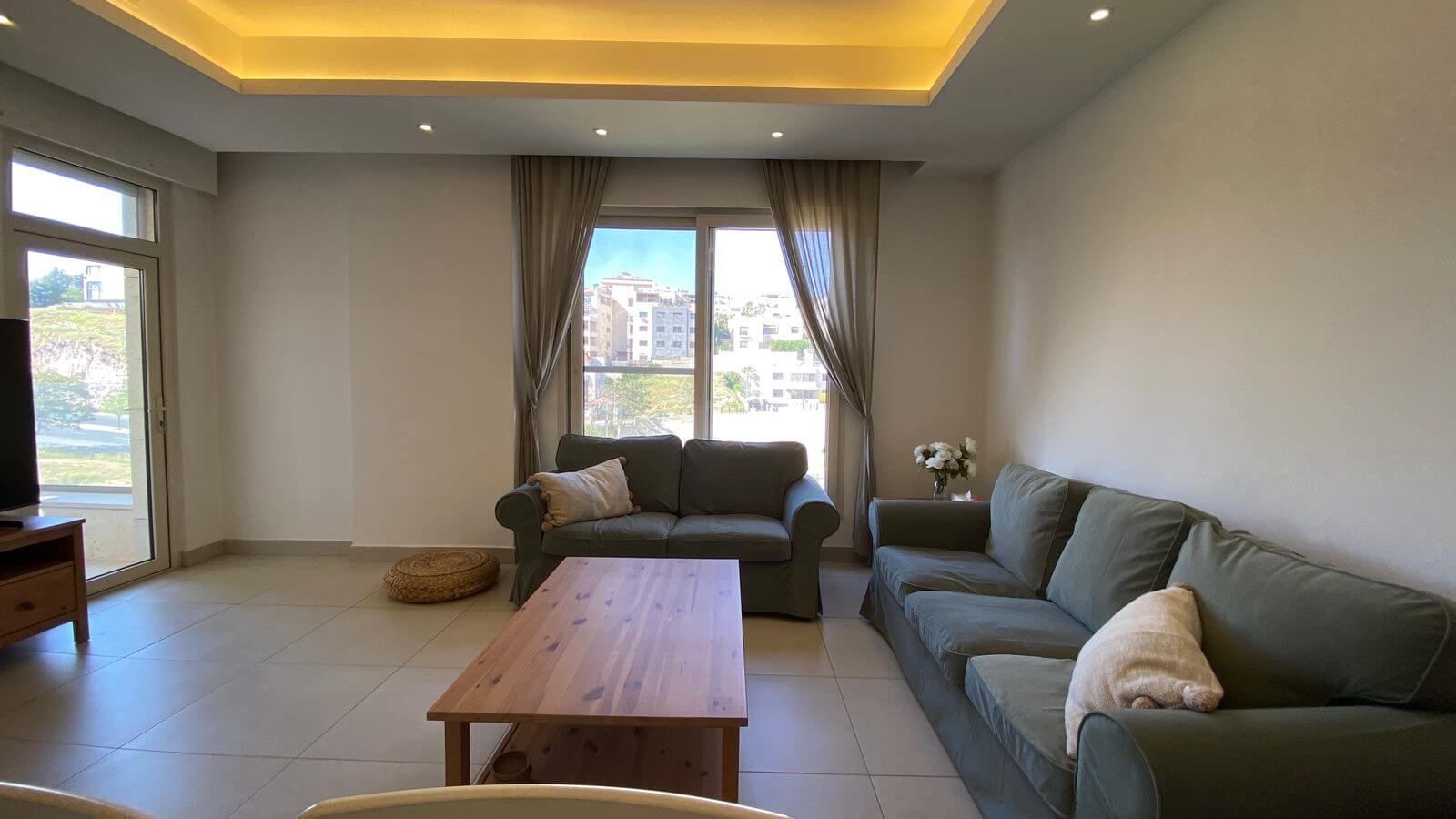 a9a2cabc-ff76-4145-855e-dc87f7d54ace Living room with two gray sofas, a wooden coffee table, and a TV on the left; large windows with beige curtains.