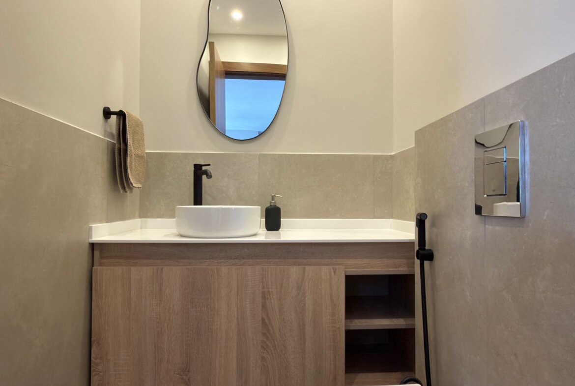 Modern bathroom sink area with a white round vessel sink, black faucet, soap pump, and wood vanity beneath beige tiled walls with an oval mirror above.