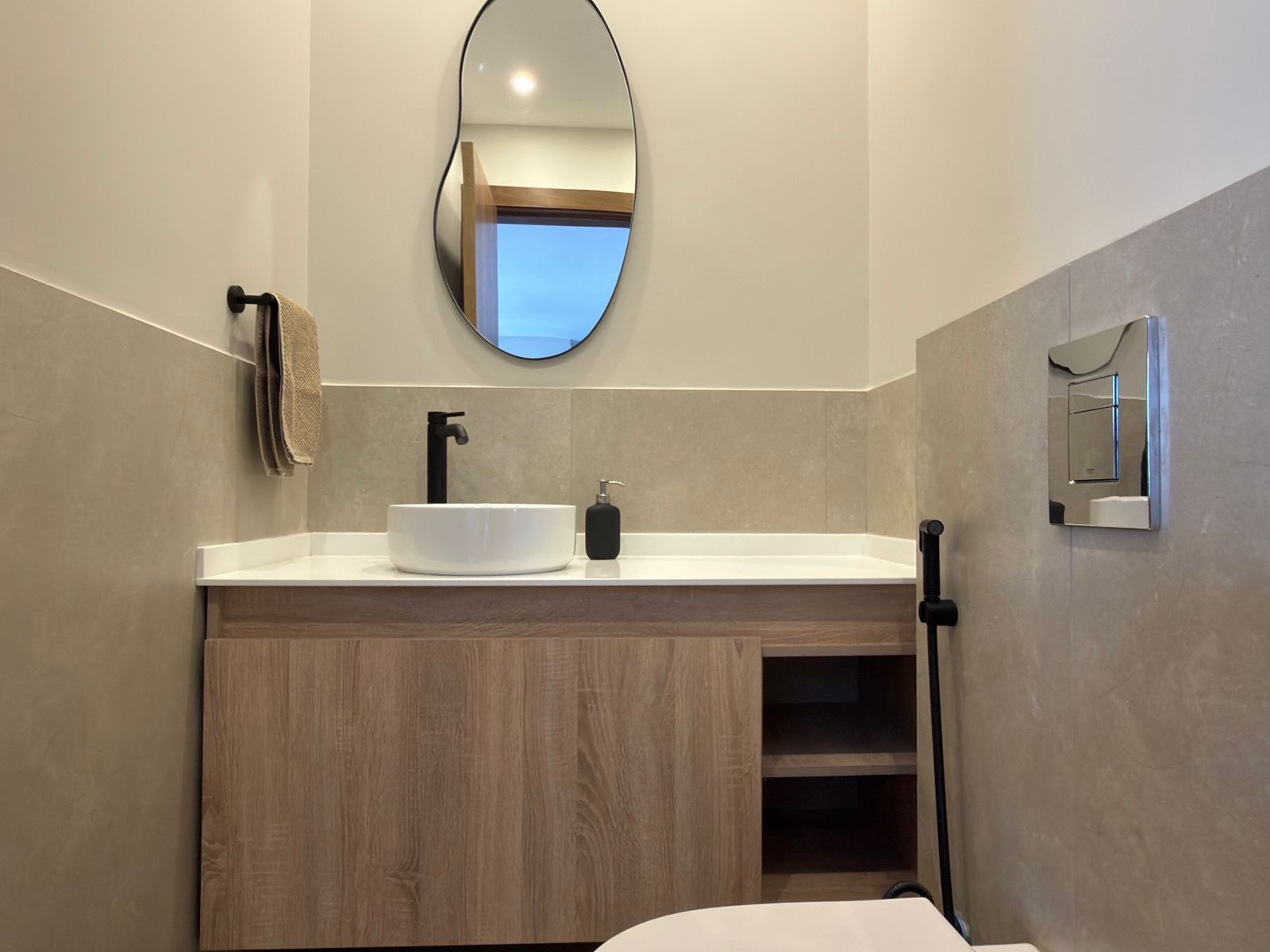 a9a3ad58-e22b-4ced-92d9-699524278c49 Modern bathroom sink area with a white round vessel sink, black faucet, soap pump, and wood vanity beneath beige tiled walls with an oval mirror above.