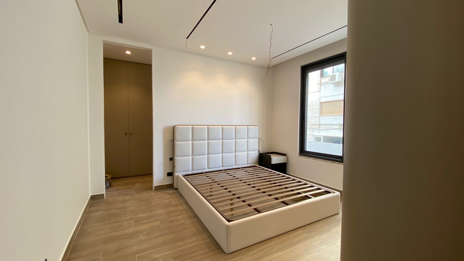 a9f42630-87a0-4727-b9d2-2fe9d564bd08 Modern minimalist bedroom with a white upholstered bed frame and slatted foundation, two dark nightstands, and a large window; ceiling wires and recessed lights indicate ongoing setup.