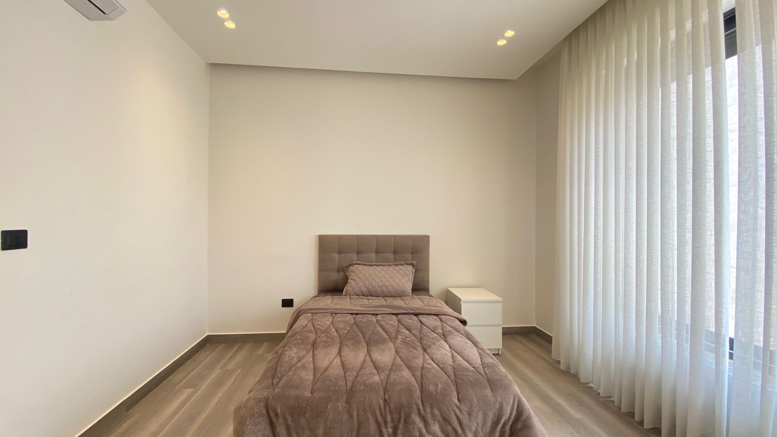 aa31eab8-5a85-4416-8329-dbe5731dade2 Single bed with tufted headboard and taupe bedding in a minimalist room, white walls, and a white nightstand nearby.