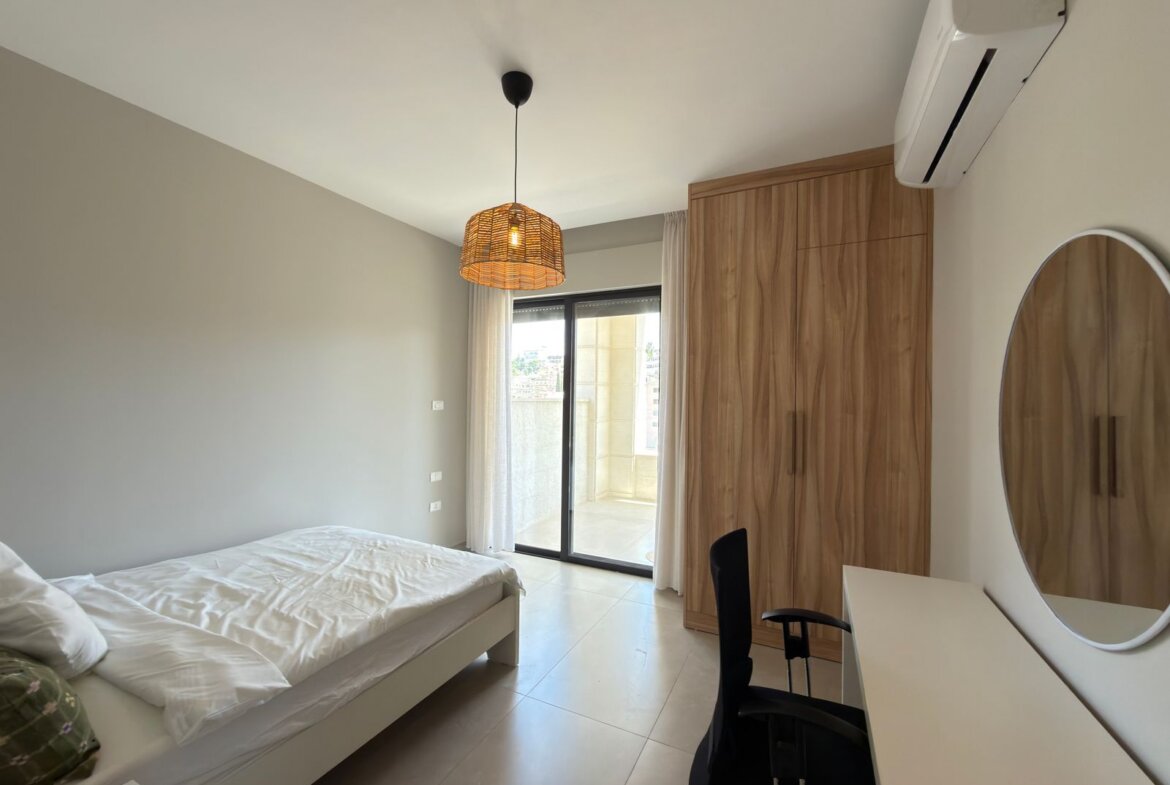 Modern bedroom with a low white bed, tall wooden wardrobe, desk and round mirror; glass balcony door with sheer curtains nearby.