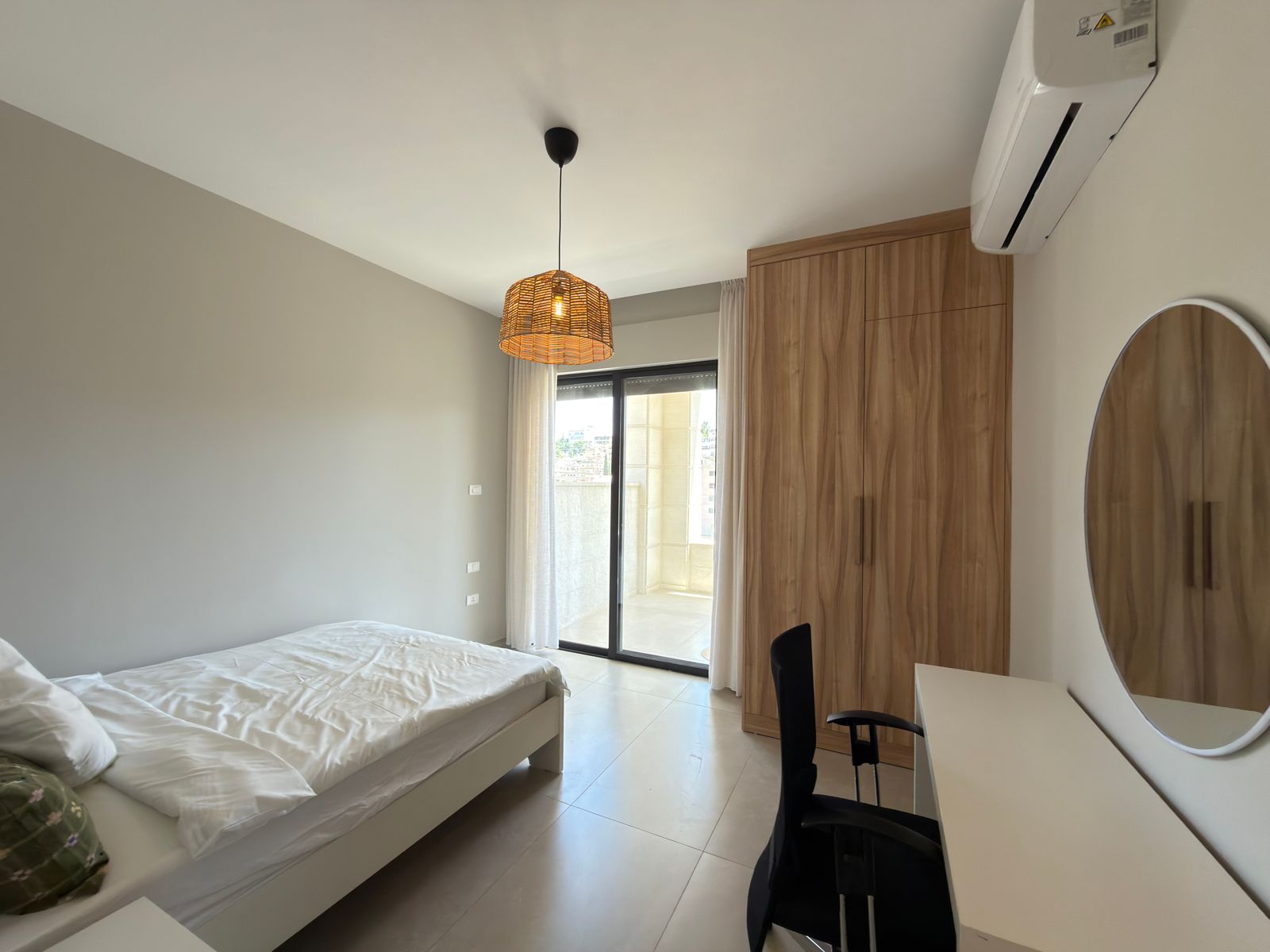 aa599b2e-76c3-4710-affa-87110cbaa25e Modern bedroom with a low white bed, tall wooden wardrobe, desk and round mirror; glass balcony door with sheer curtains nearby.