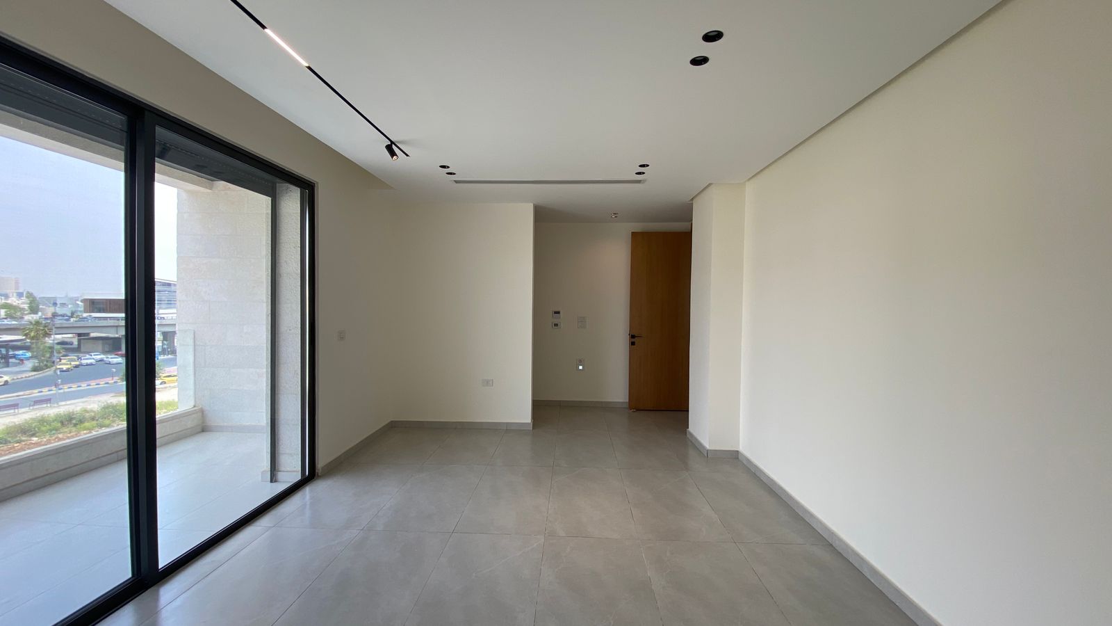 aa87152d-32ed-4dc4-a729-886ae1913917 Empty modern living room with large glass sliding doors to a balcony, light walls, and gray tiled floor; wood entry door on far wall.