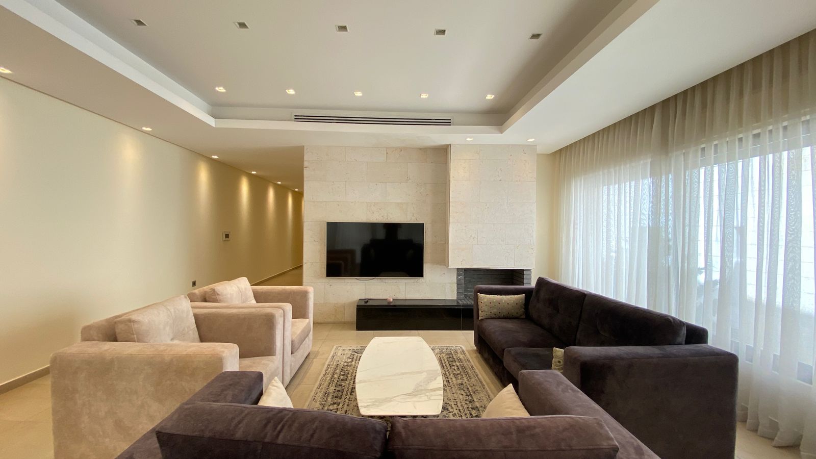 ab674781-bc97-4f16-b14a-12e548041d71 Contemporary living room with beige and brown sofas, a white marble coffee table, TV on a stone wall, and sheer-curtained windows.