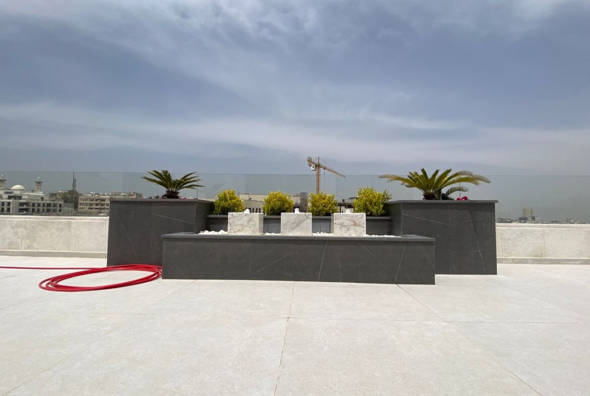 Rooftop terrace with dark stone planters and small shrubs, palm trees, a red hose on light tiles, and a distant city skyline with a crane under a cloudy sky.