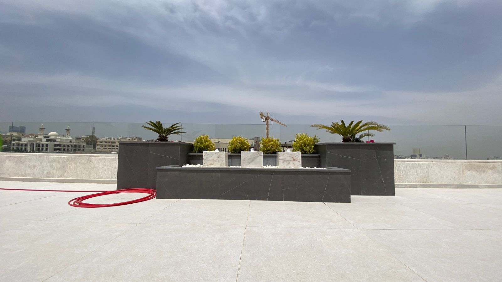 ac74b316-dbfe-4daf-9b64-903623039e79 Rooftop terrace with dark stone planters and small shrubs, palm trees, a red hose on light tiles, and a distant city skyline with a crane under a cloudy sky.