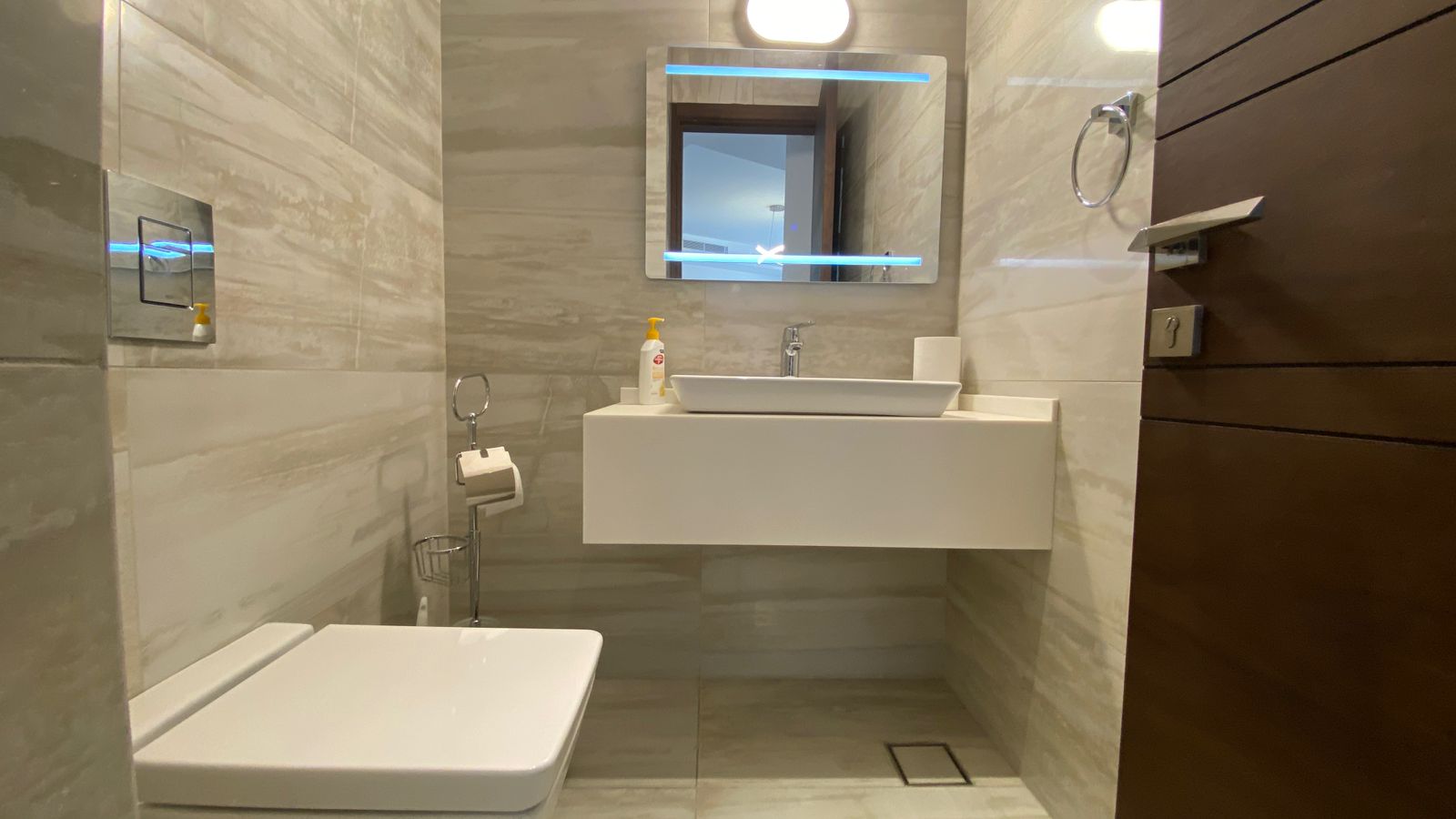 ad33604e-a441-4e21-a19f-6c84852eb0be Modern bathroom with a floating white sink, backlit mirror, soap bottle, and wall-mounted toilet. The wooden door is open to the room.