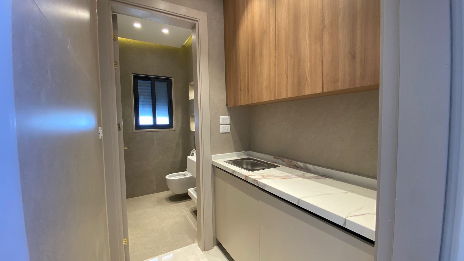 ad5dfe08-45cd-4dfe-95da-bfd8e3945043 Compact kitchen with white marble countertop and sink, wood upper cabinets, opening to a gray-tiled bathroom with toilet and bidet visible in the doorway.
