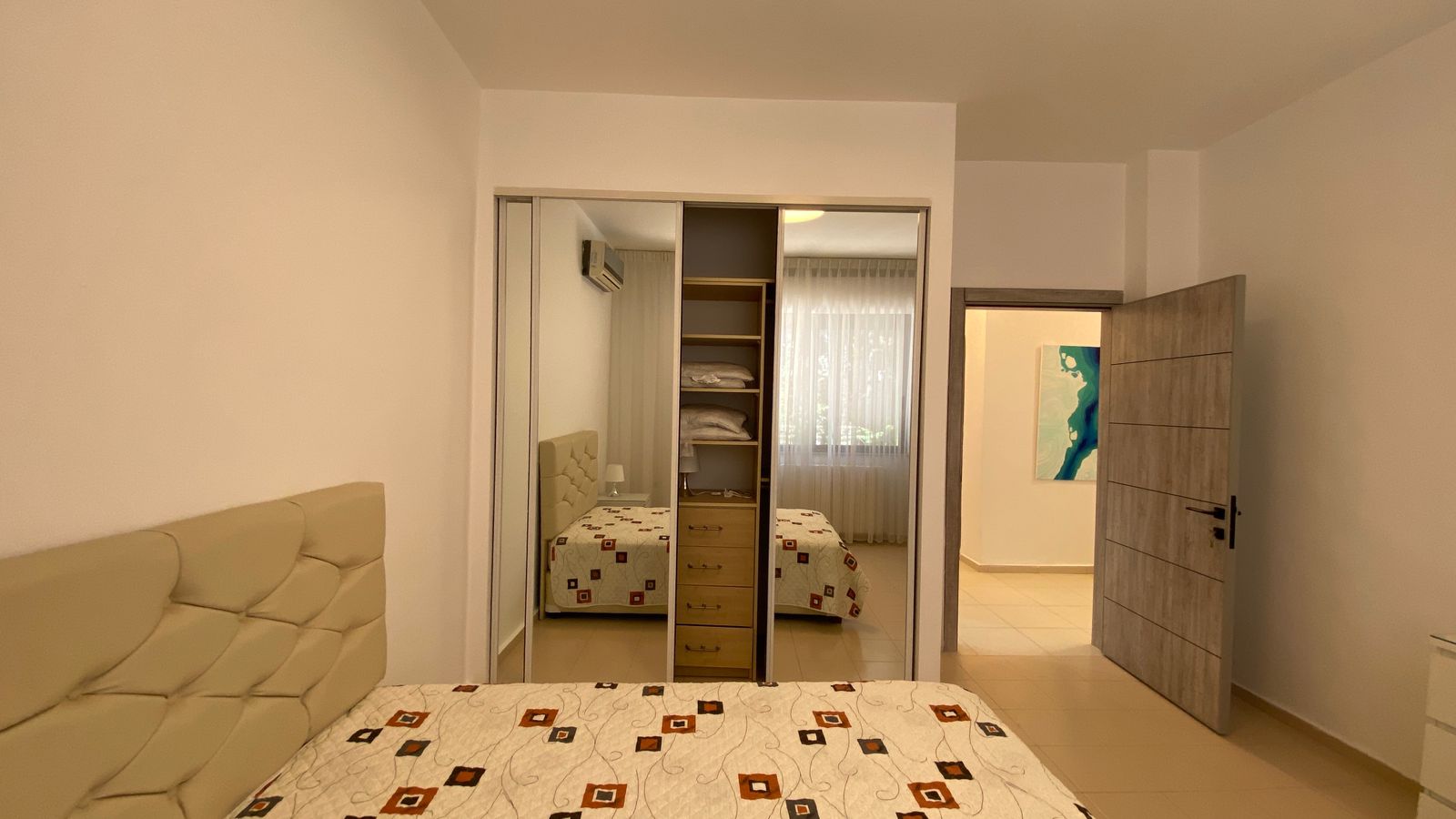 ad5ed08b-792a-484a-aef3-2a28173a370c Bedroom with a padded beige headboard, patterned quilt, and a mirrored wardrobe reflecting the bed. Open door leads to a bright hallway with a blue-green artwork nearby.