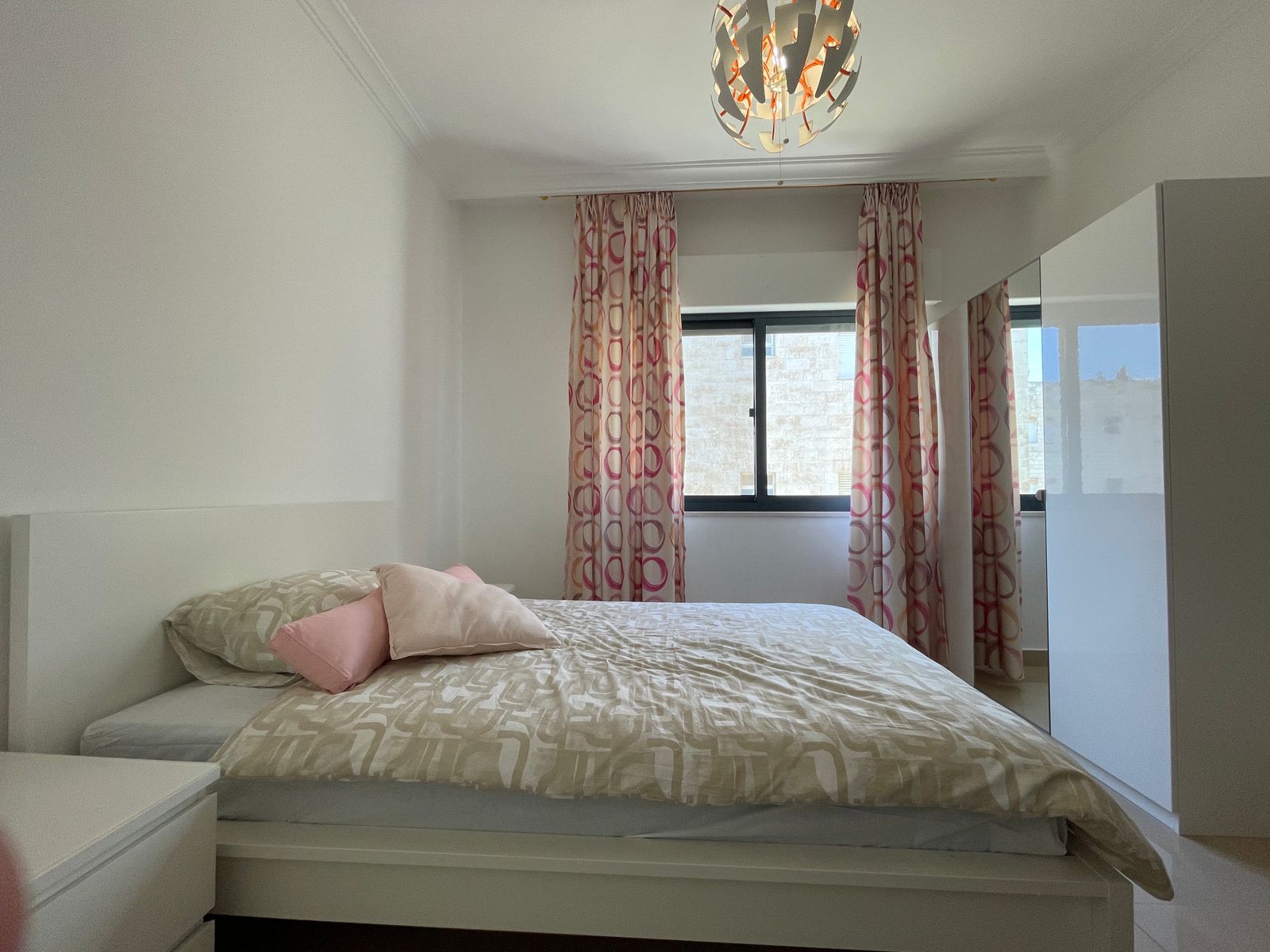 ad7ae715-a650-4302-a48b-ade98c528eb4 Bright white bedroom with a low platform bed, beige geometric duvet, pink pillows, and a white wardrobe with mirrored doors.