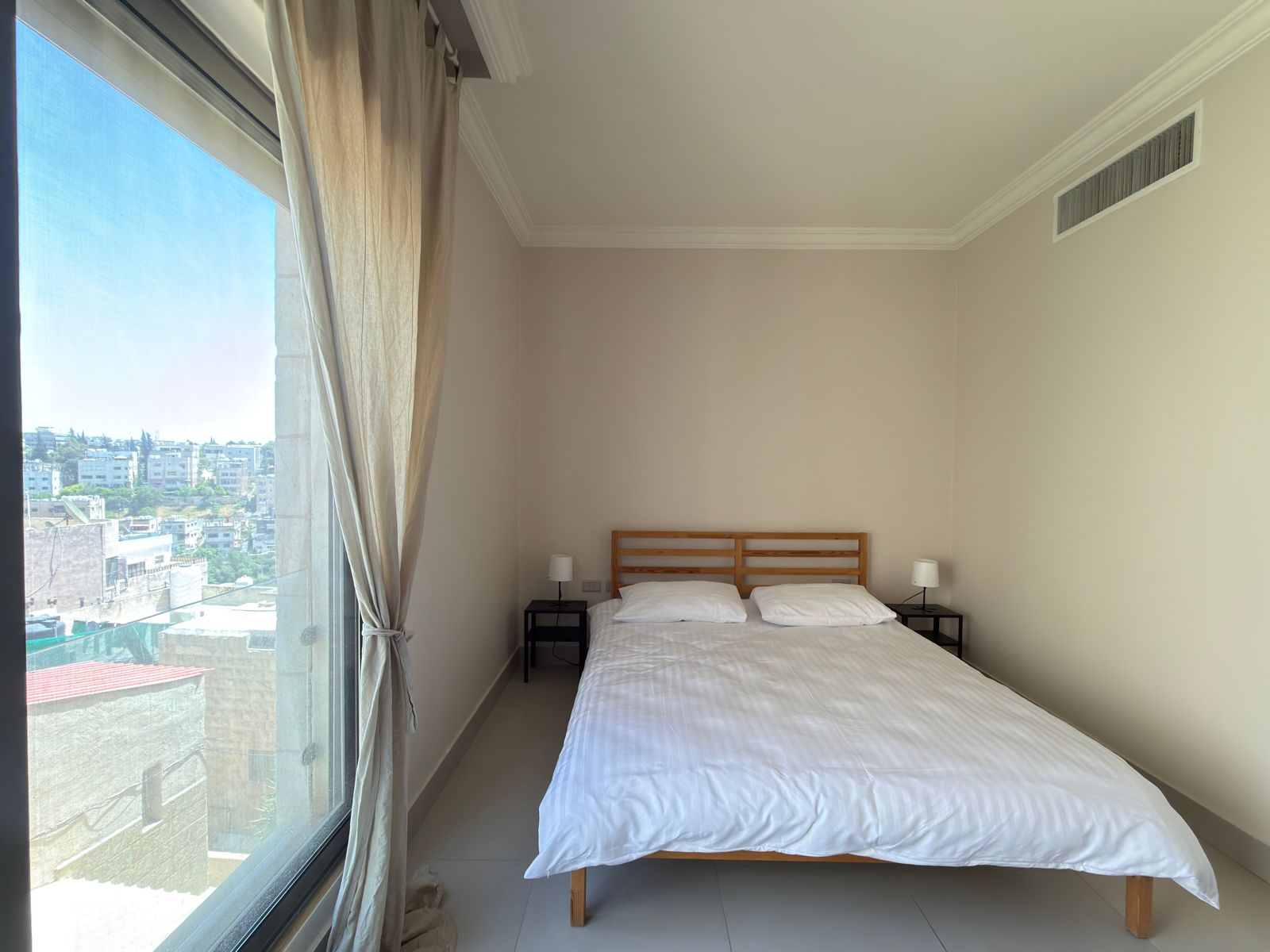 ad7fed19-1406-4606-b992-b25b2092c9fc Bedroom with a wooden-framed bed, white linens, two black nightstands, and matching lamps, beside a large window with a city view.