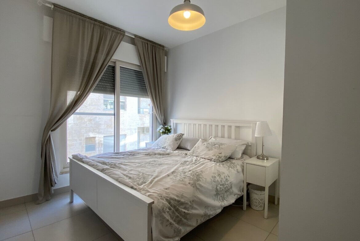 ada4459f-54d2-4c6d-b38f-0c1049de82cb Bright white bedroom with a white bed, floral bedding, and a nightstand lamp by a large window.