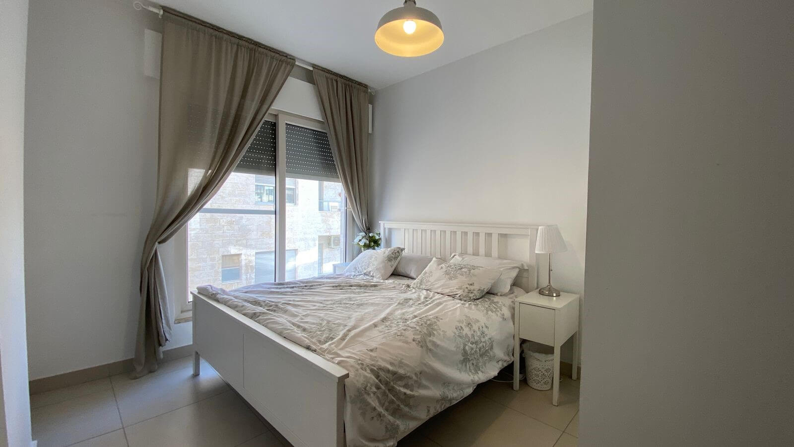 ada4459f-54d2-4c6d-b38f-0c1049de82cb Bright white bedroom with a white bed, floral bedding, and a nightstand lamp by a large window.