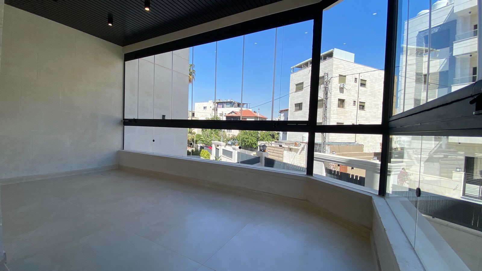 adb33289-759d-4b35-8847-61e24deb0ed8 Empty bright room with large floor-to-ceiling windows overlooking neighboring buildings on a sunny day