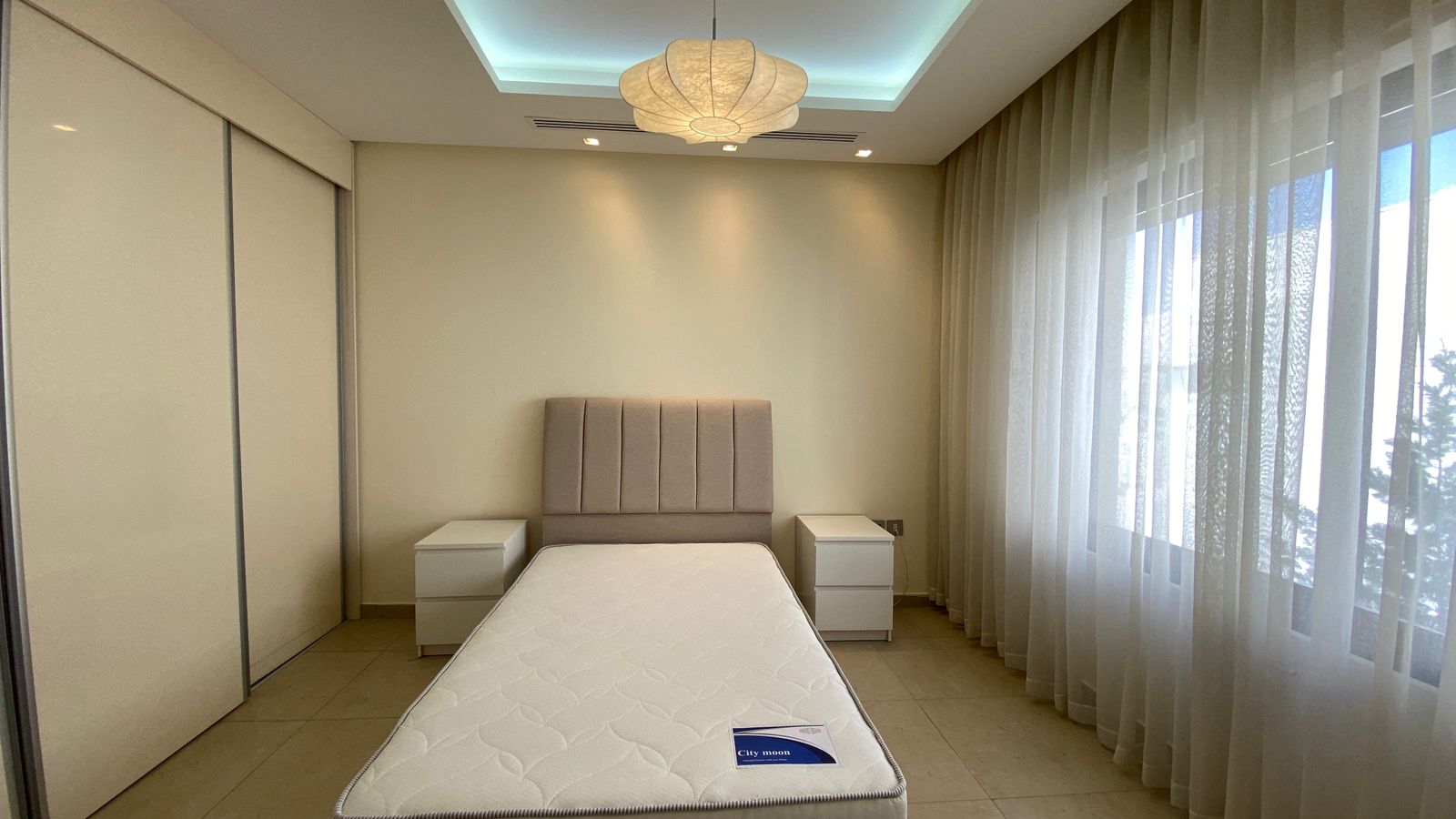 adea4944-8dcf-4454-8db5-dd4ae49cb963 Minimalist bedroom with a padded headboard bed, two white nightstands, and sheer curtains along large windows.
