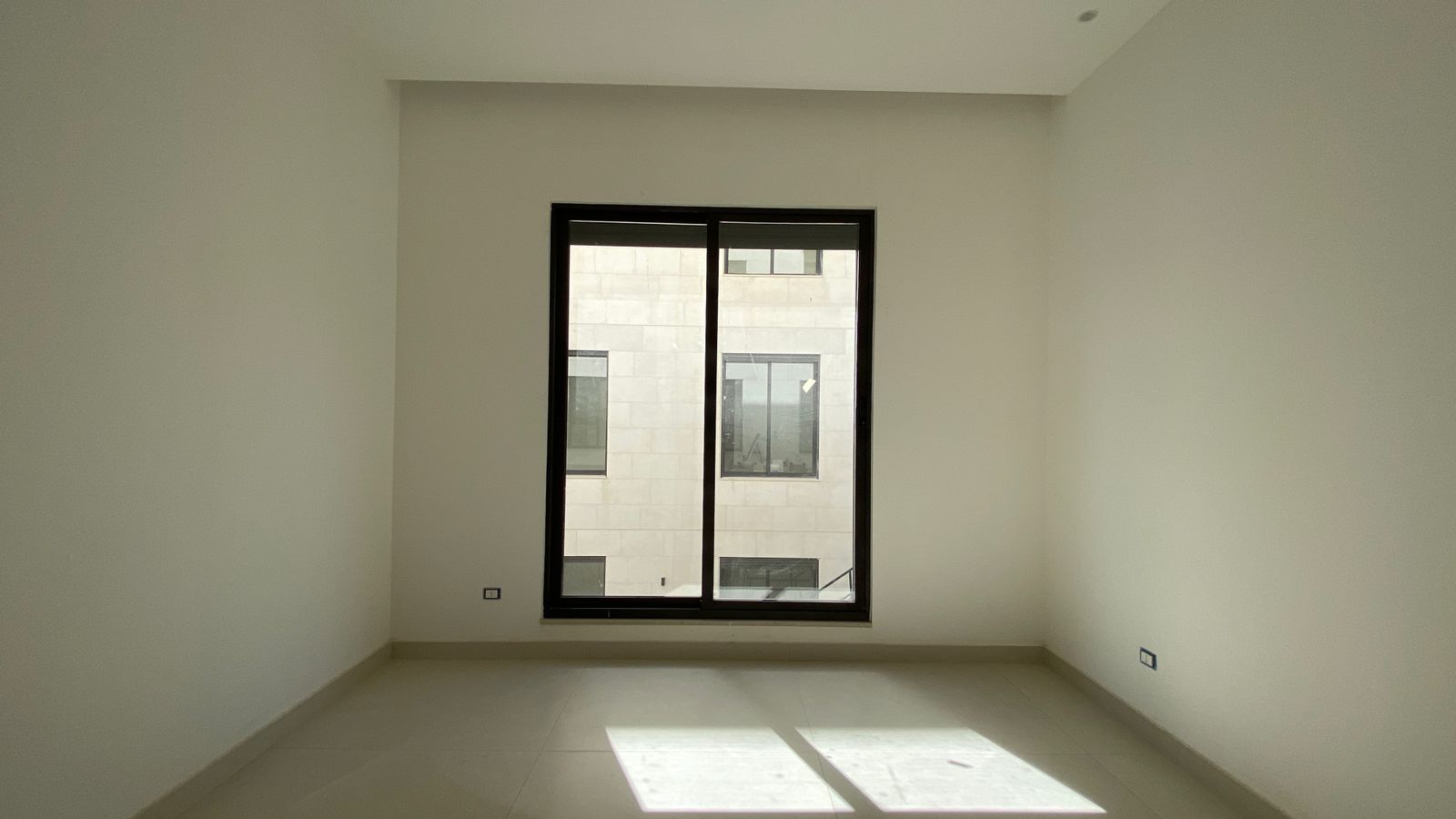 ae6e4acc-aa2b-4bee-a738-6c89a7fd4530 Empty white room with a large black-framed window, sunlight on the beige tiled floor, minimal interior.