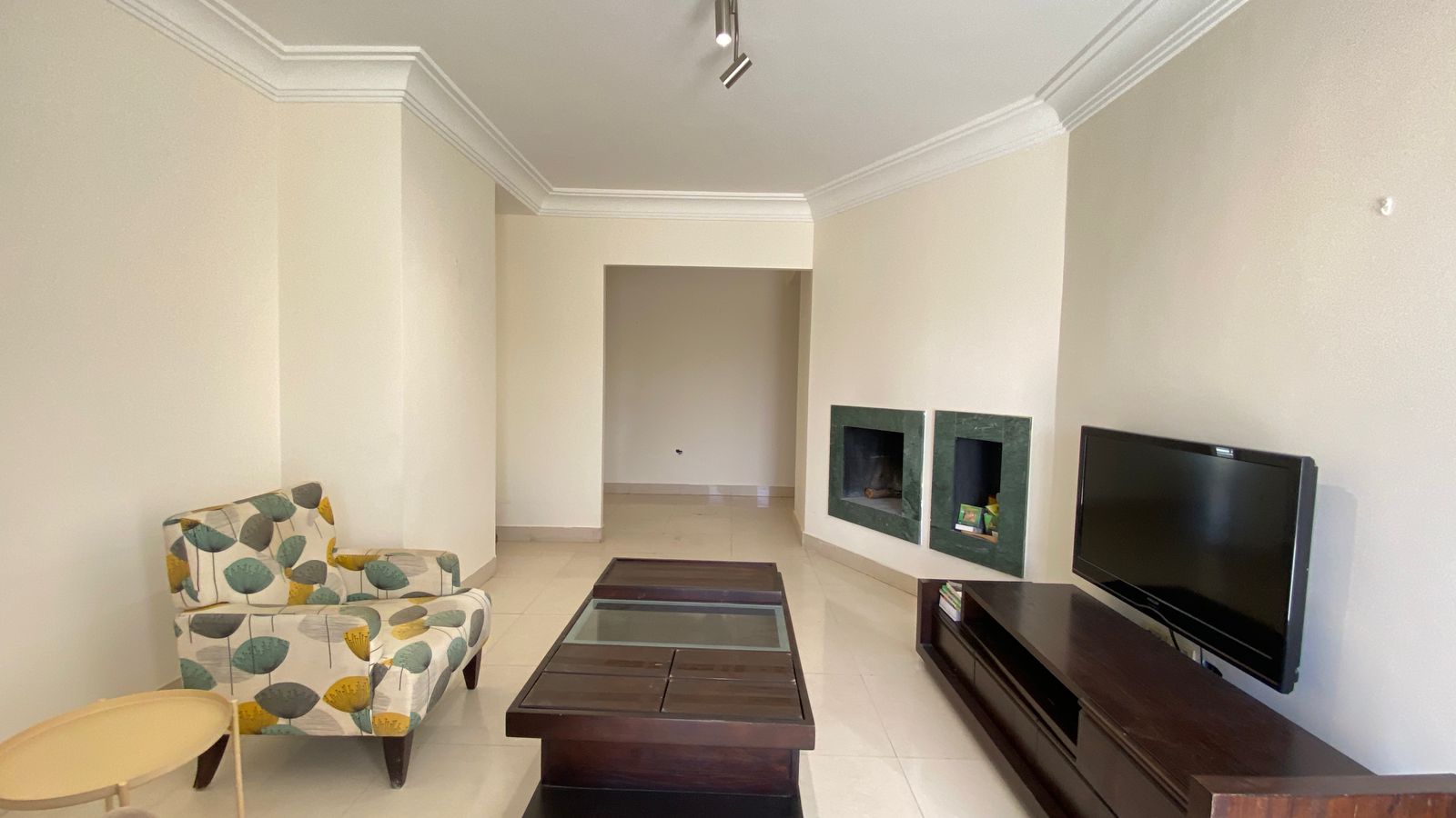 aed48357-2984-49ec-85d1-5e121273f5ae Living room with beige walls, crown molding and track lighting; a patterned armchair, glass-topped coffee table, green-framed fireplace niches, and a flat-screen TV on a dark wooden stand.