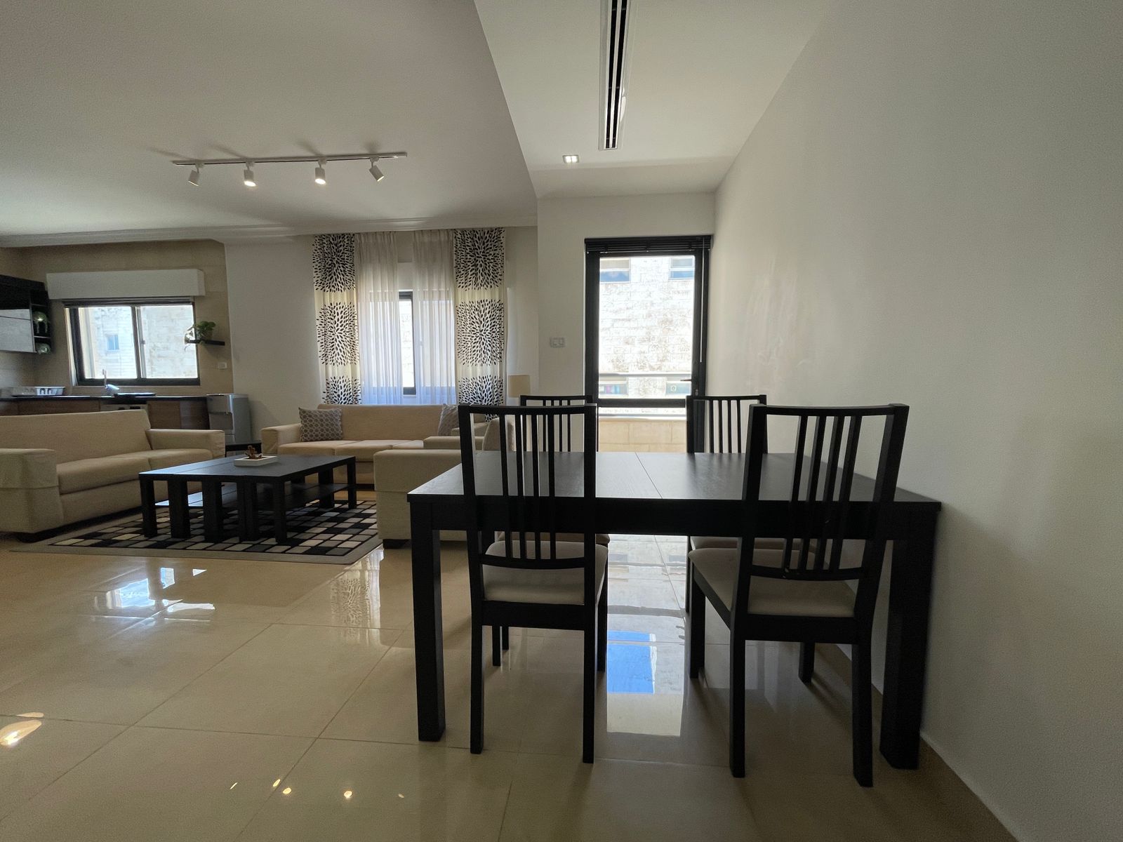 af47c701-4f67-449a-97a5-00122b8b19f9 Open-plan living and dining area with a dark rectangular dining table and black chairs, beige sofas, and large windows with sheer curtains.