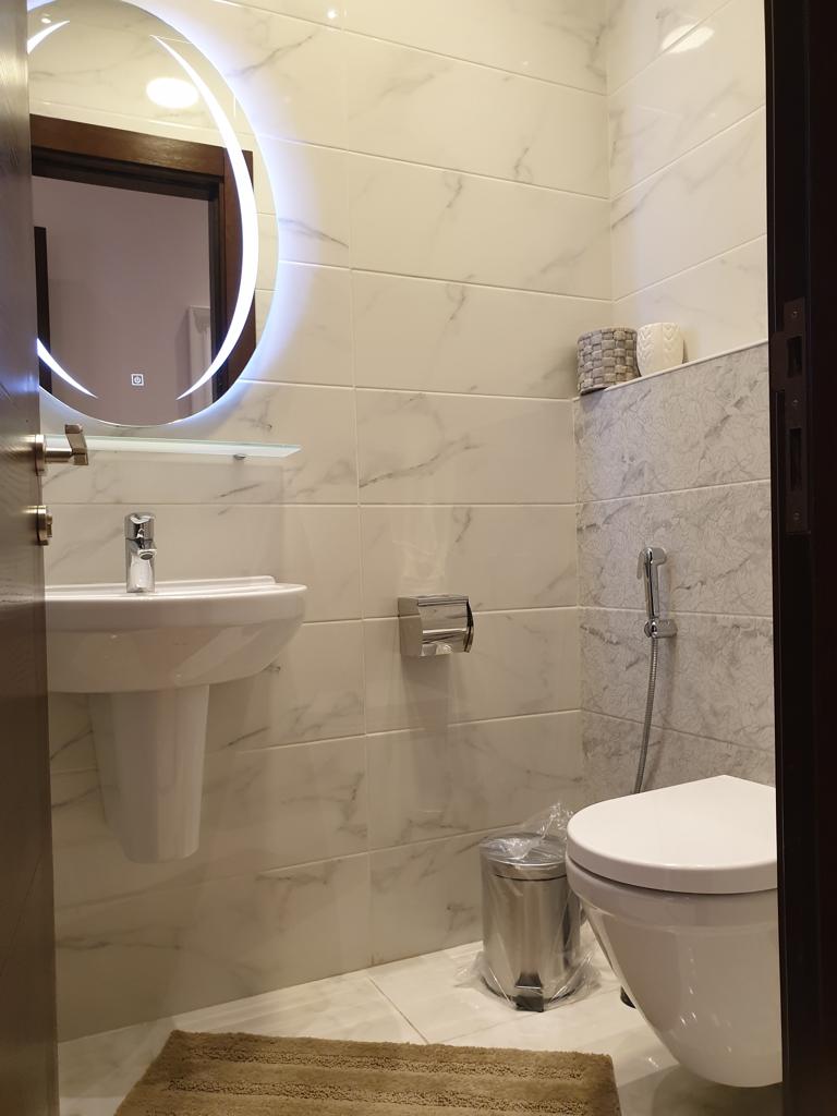 af88a251-52a8-43ca-af20-e2c9126d0df5 Circular backlit mirror above a pedestal sink in a modern marble-tiled bathroom, with a toilet and a bidet spray nearby, a small trash can, and a beige bath mat on the floor.