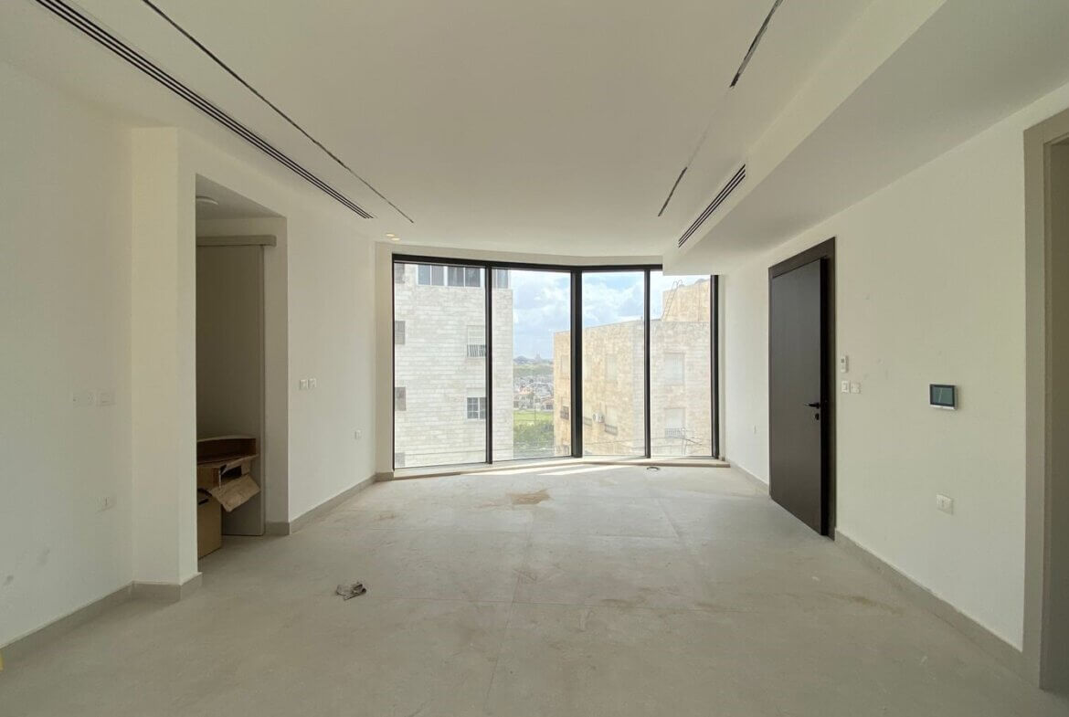 Unfurnished white living room with large curved floor-to-ceiling windows and city view, neutral tiled floor, doorway on the right.