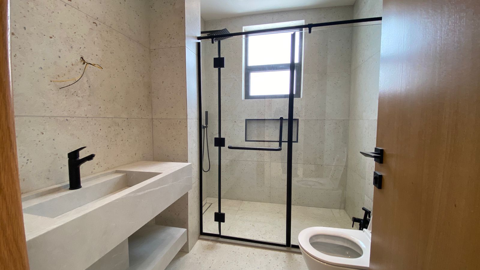 b107692e-0e8e-43c9-9d8d-538dc338b8bd Modern bathroom with a glass-framed shower, black hardware, and a concrete sink seen from the doorway