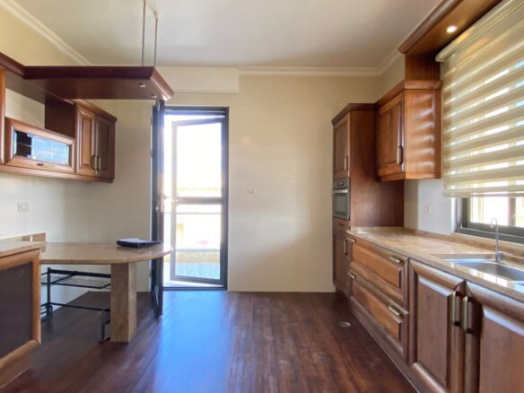 Brightwood-toned kitchen with dark wood cabinets, marble countertops, and a doorway to an outdoor area; large window with horizontal blinds on the right.