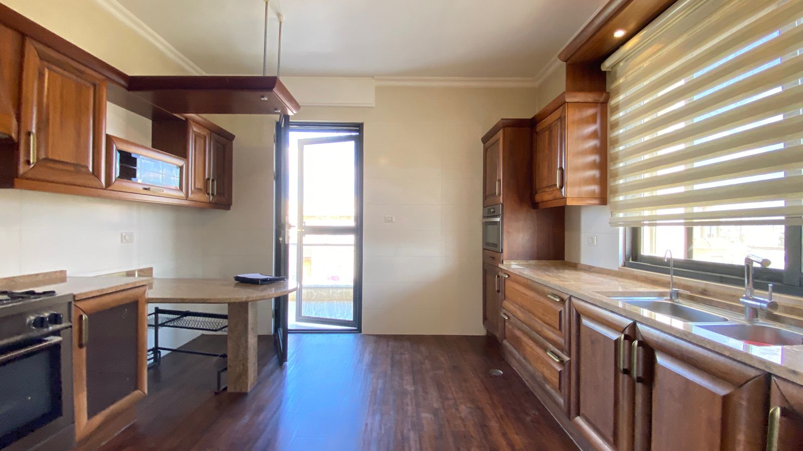 b1c1b57a-293e-4f40-b85d-2700668c4e47 Brightwood-toned kitchen with dark wood cabinets, marble countertops, and a doorway to an outdoor area; large window with horizontal blinds on the right.