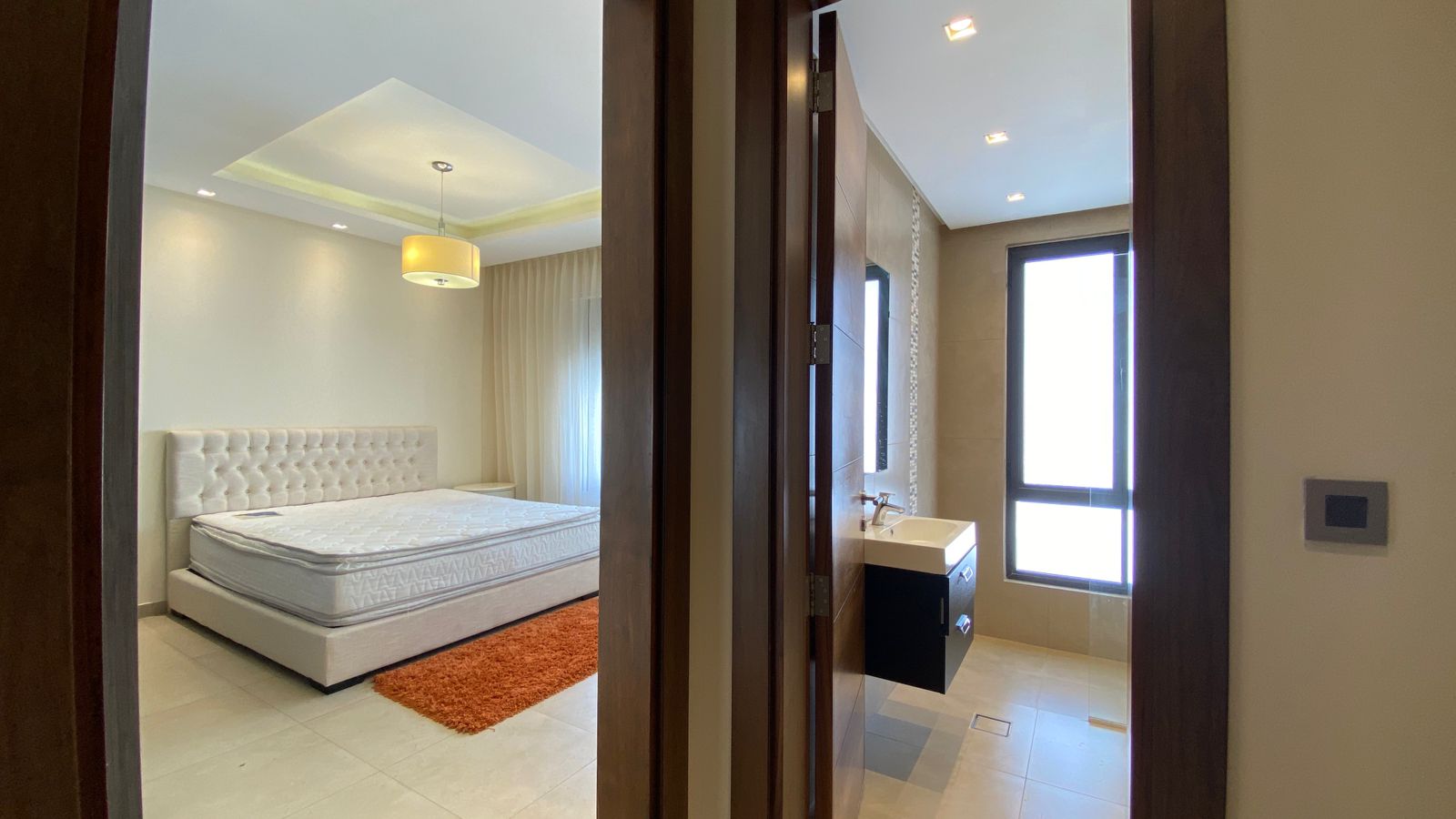 b1dd389d-09ad-4721-8c21-482f98d28da7 Modern bedroom with a white tufted headboard bed, cream walls, and an orange shag rug; doorway opens to a bright en-suite bathroom.