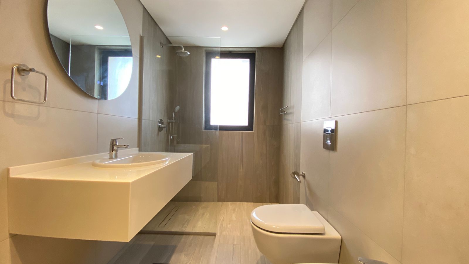 b1fde5df-e2fe-4fb6-a752-9e46e3310f75 Modern bathroom with wall-mounted sink, circular mirror, toilet, and glass-enclosed shower opposite a window.