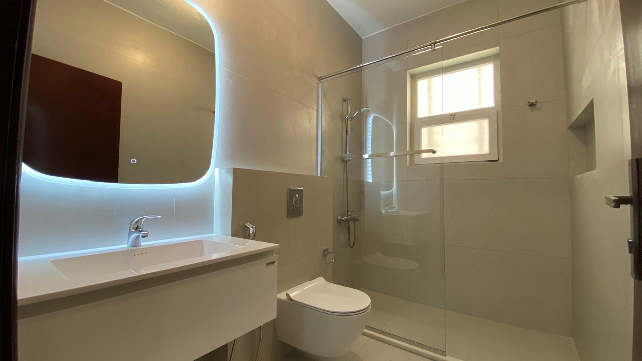b24aaf1c-4a2e-4a9e-b590-4e5df6024f2f Modern bathroom with glass shower enclosure, wall-mounted toilet, and white vanity beneath a backlit oval mirror.