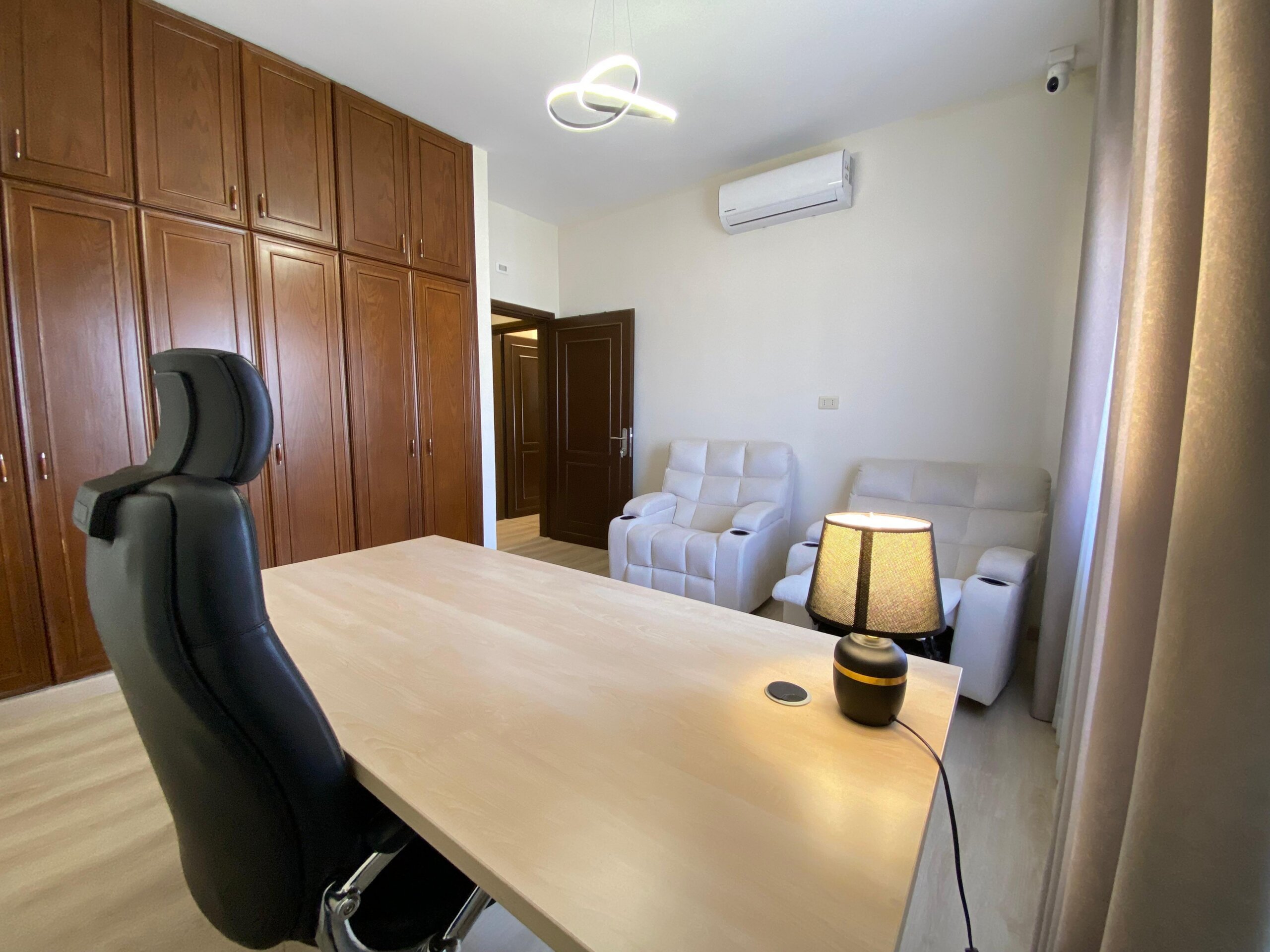 b289412c-7686-4a16-a841-6f0143054812 Home office: wooden floor-to-ceiling cabinets on left, light wood desk, black ergonomic chair, and a small seating area with white tufted chairs in back.