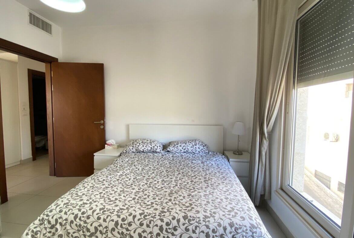 b4083283-11d8-4f21-b9a3-1d6e2cd8a6fe Bright modern bedroom with a double bed, white headboard, two white nightstands, and a lamp on the right; large window with beige curtains on the right side fills the room with light.